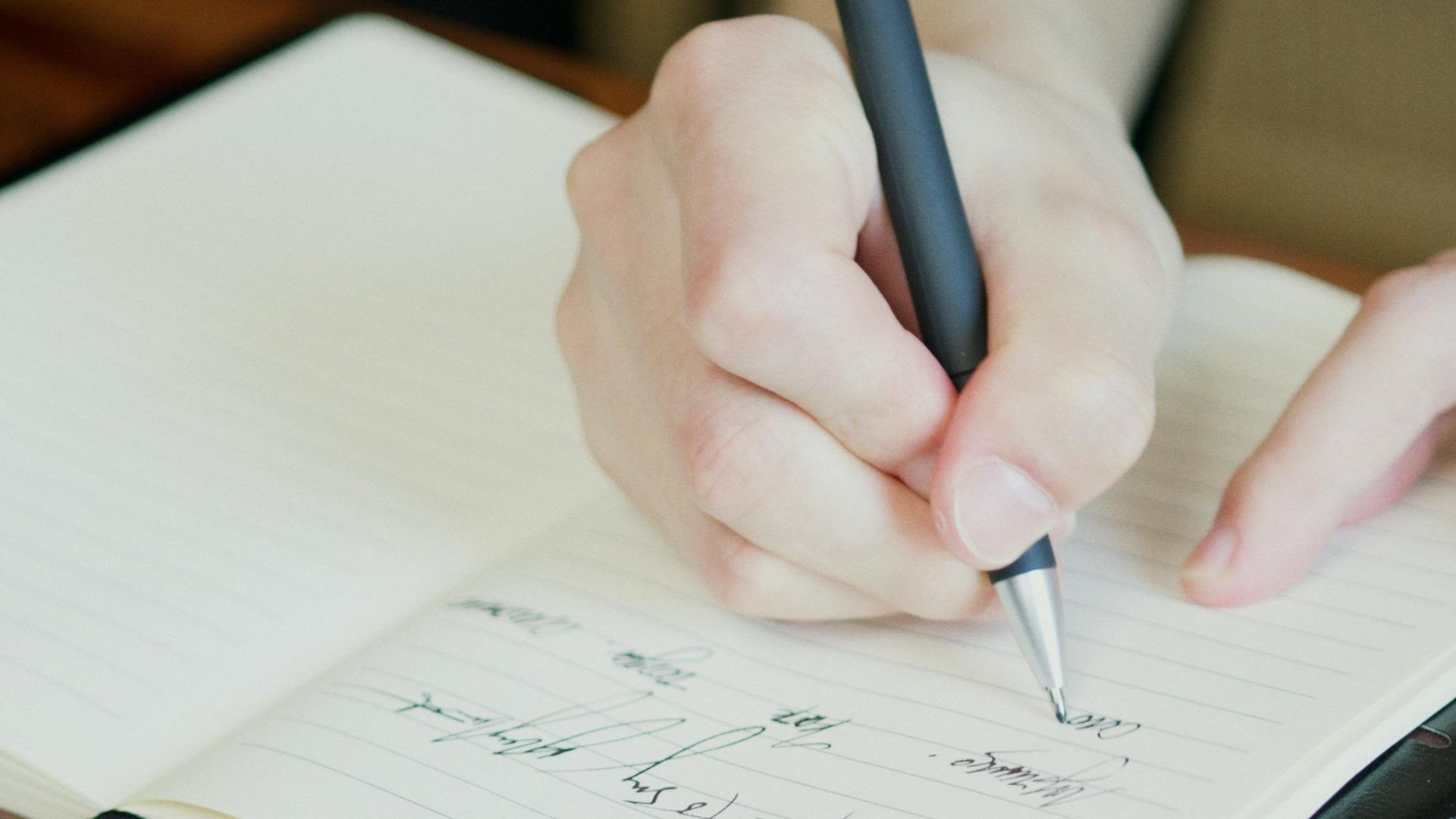 A hand holding a pen writing in an open notebook on a wooden table.
