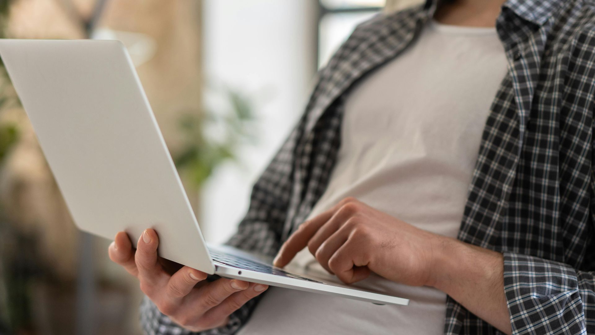 Young adult in casual shirt using a laptop indoors with blurred background and shallow focus.