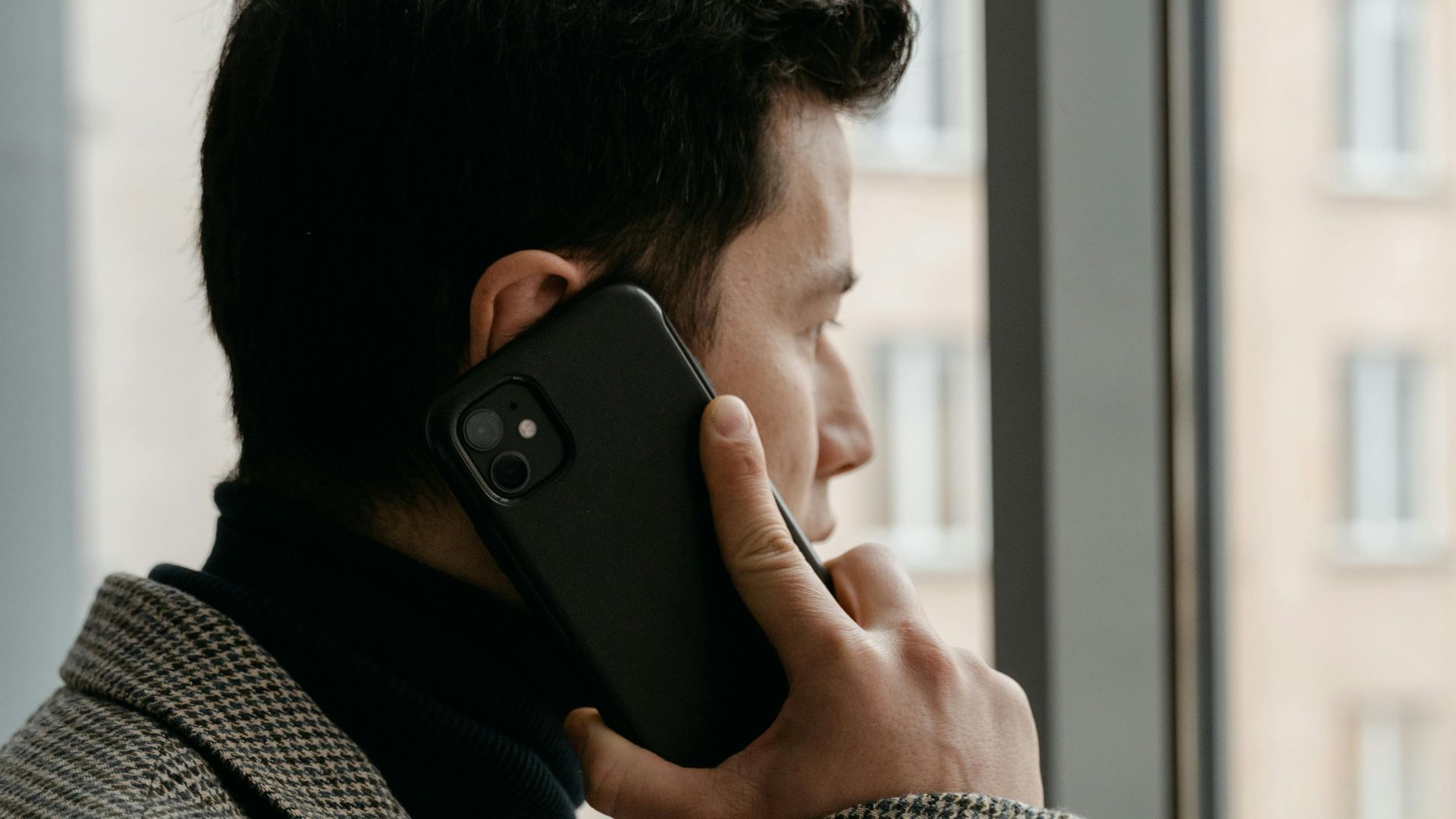 Man in a checkered blazer using a smartphone while gazing out a window.