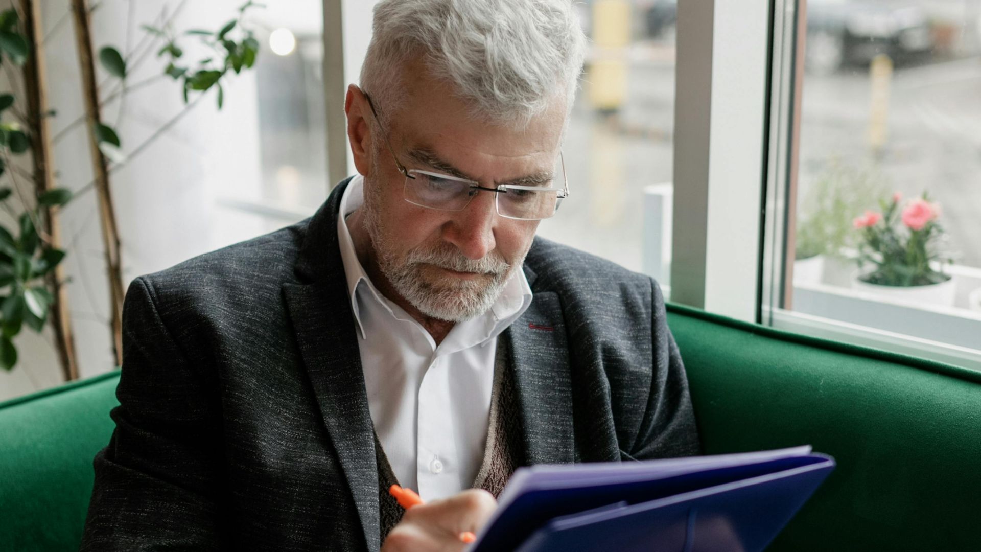 Senior man with gray hair writing on a tablet indoors, wearing glasses and a suit.