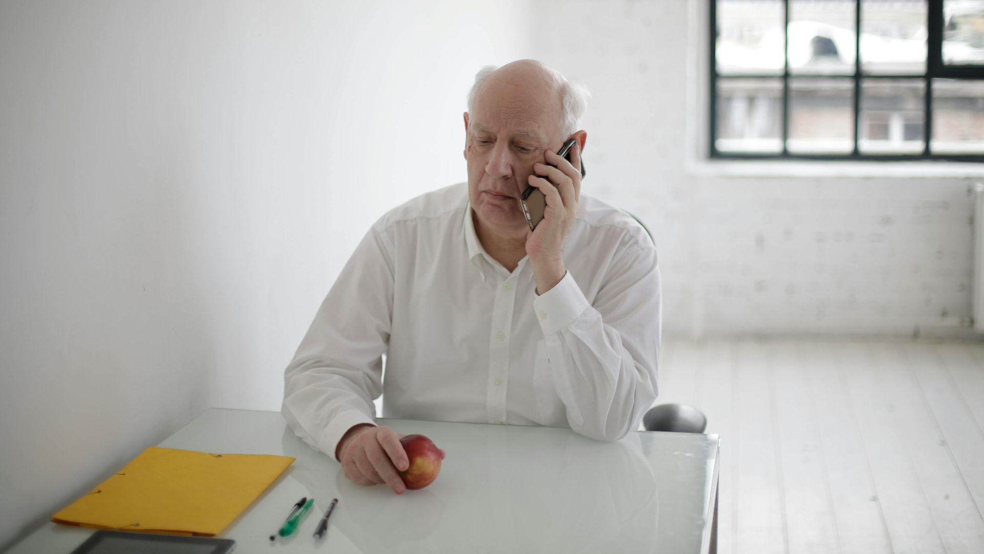 Serious male manager in formal clothes talking on phone while sitting at table with apple and documents in spacious room
