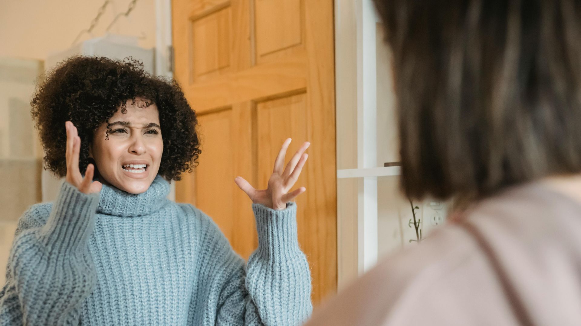 Angry young multiracial ladies in casual clothes standing in bright apartment near door while having disagreement and looking at each other
