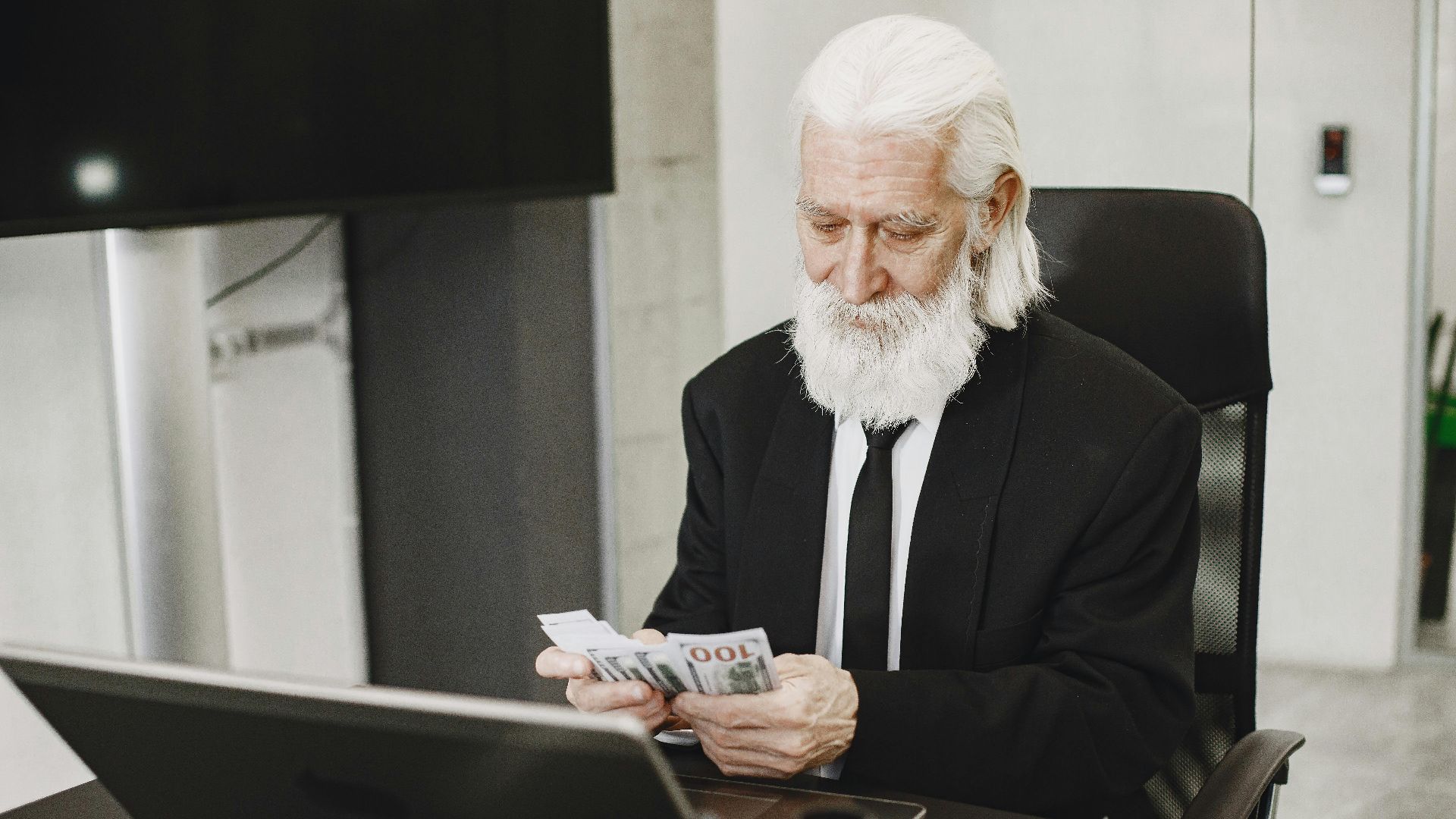 Senior businessman in suit counting money at office desk with a laptop. Elegance and financial focus in modern workspace.