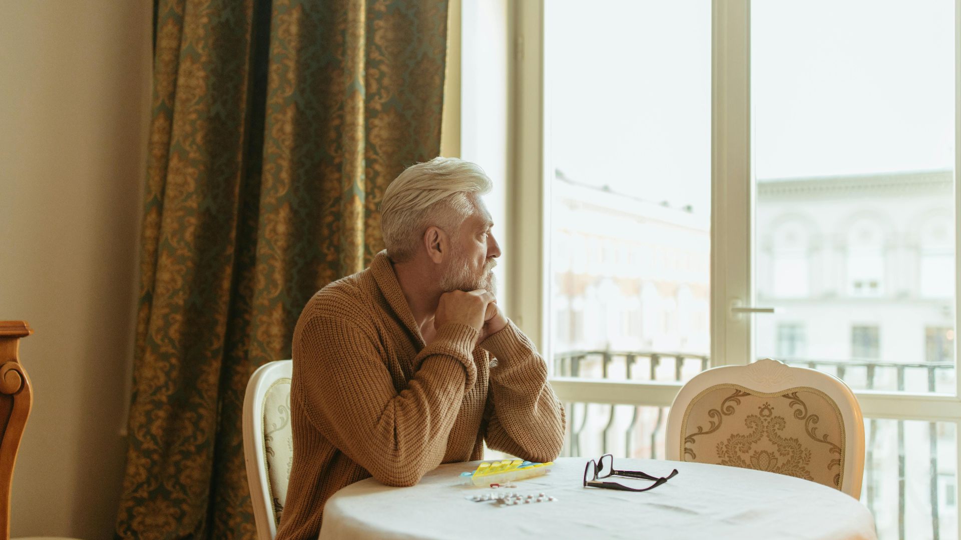 Elderly man in a cozy room gazing thoughtfully out the window during the day.