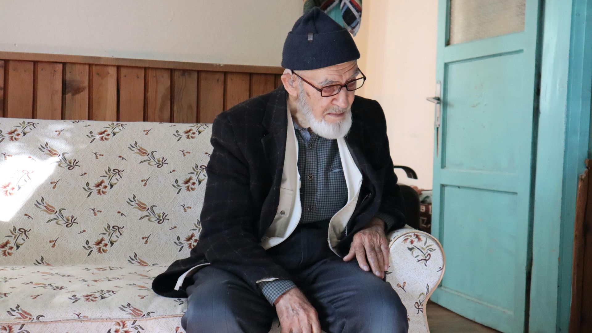 Elderly man sitting on a floral sofa indoors, enjoying a moment of relaxation and contemplation.
