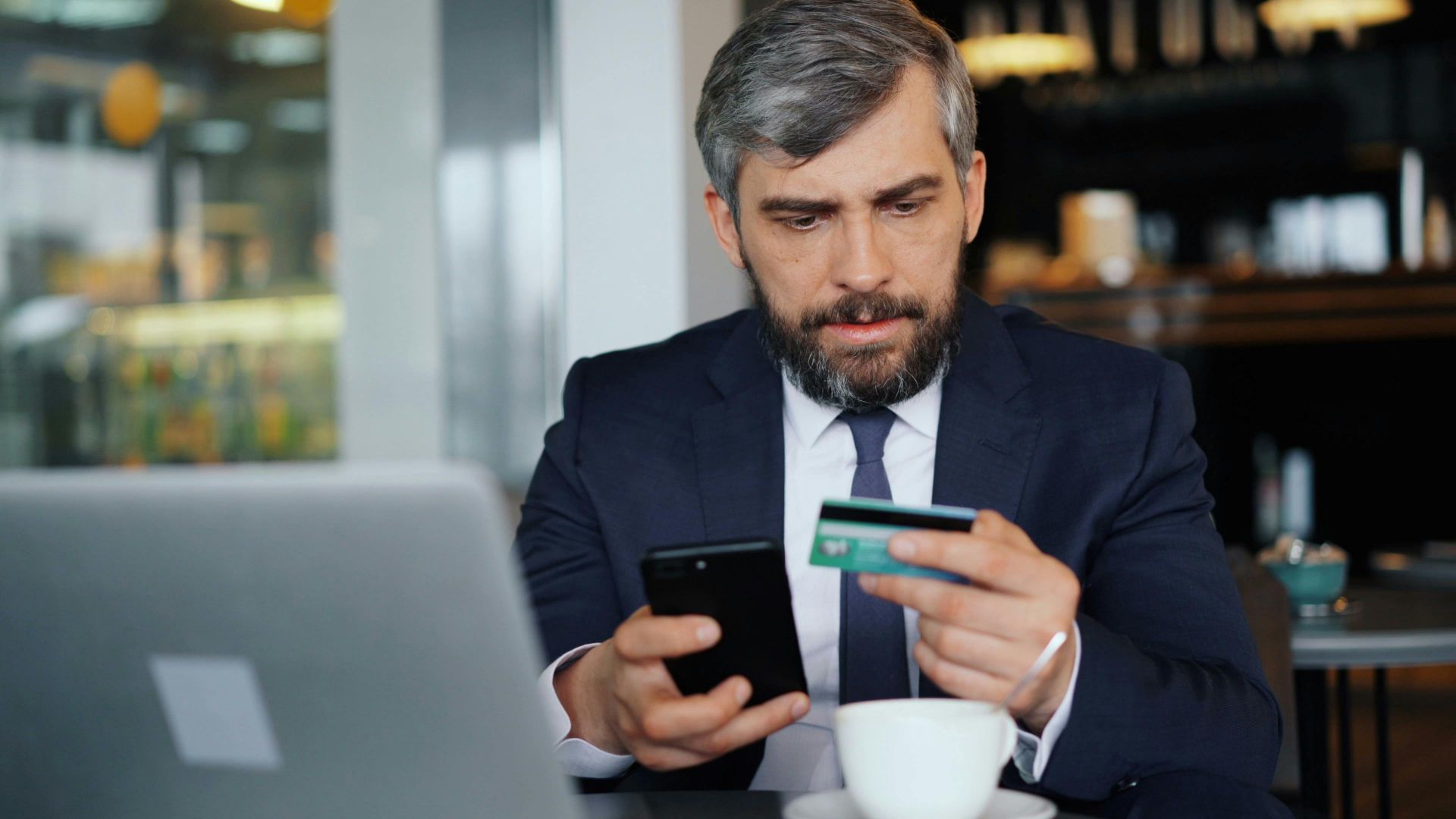 A businessman using a smartphone and credit card for online banking in a modern café.