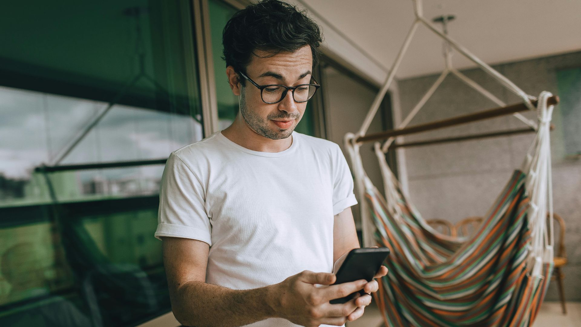 Young man in glasses using smartphone indoors with striped hammock.
