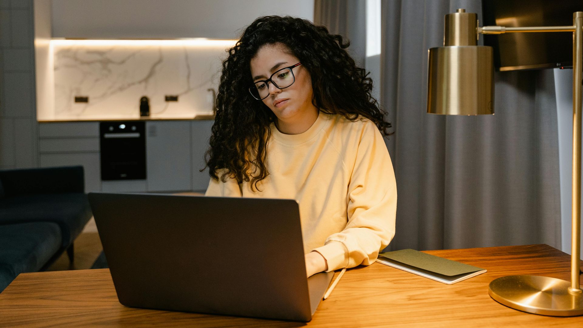 Woman in a yellow sweatshirt using a laptop at home, focusing on remote work in a cozy, modern interior.