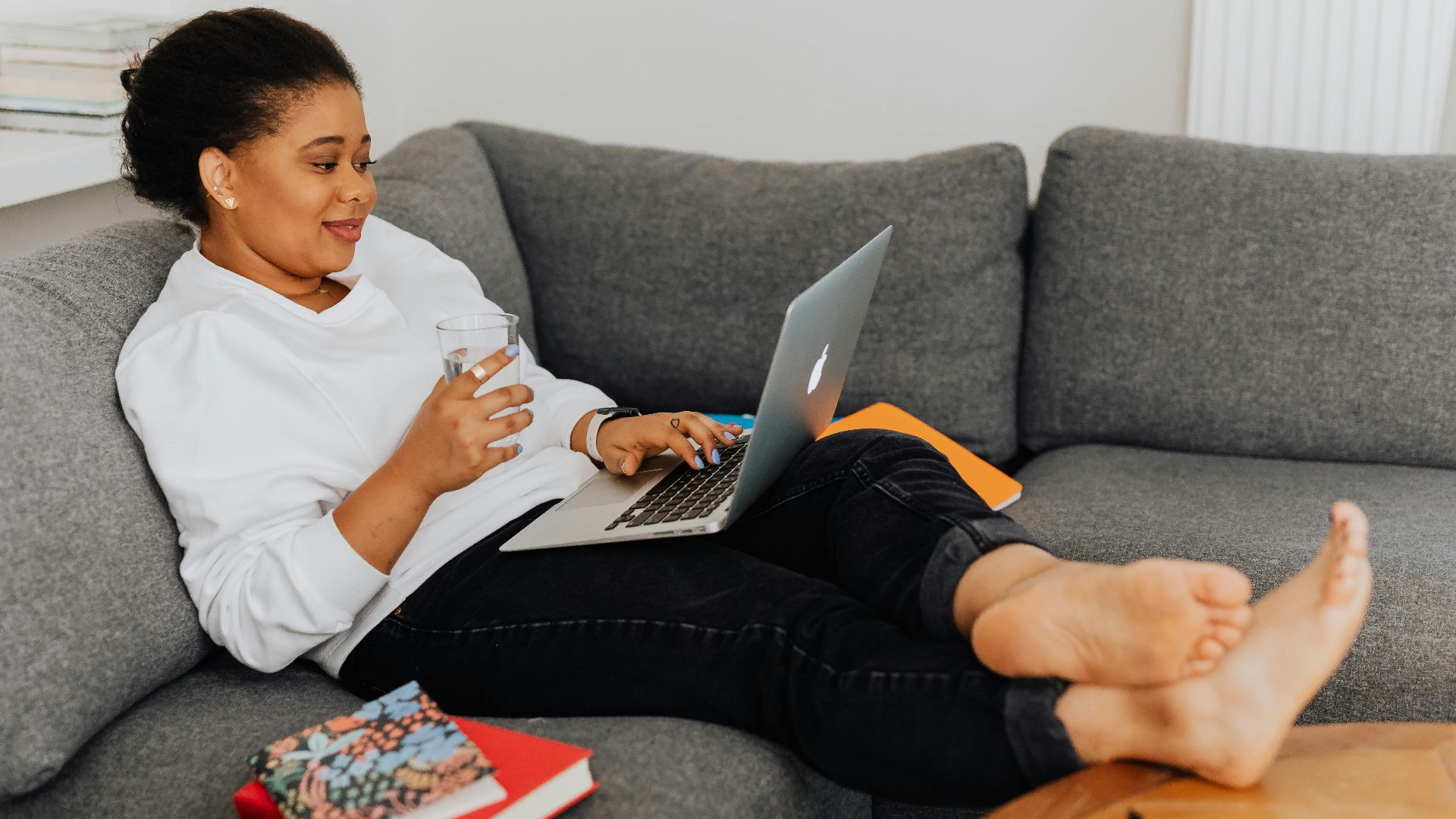 Woman comfortably lounging on a sofa, working on a laptop while holding a glass of water.