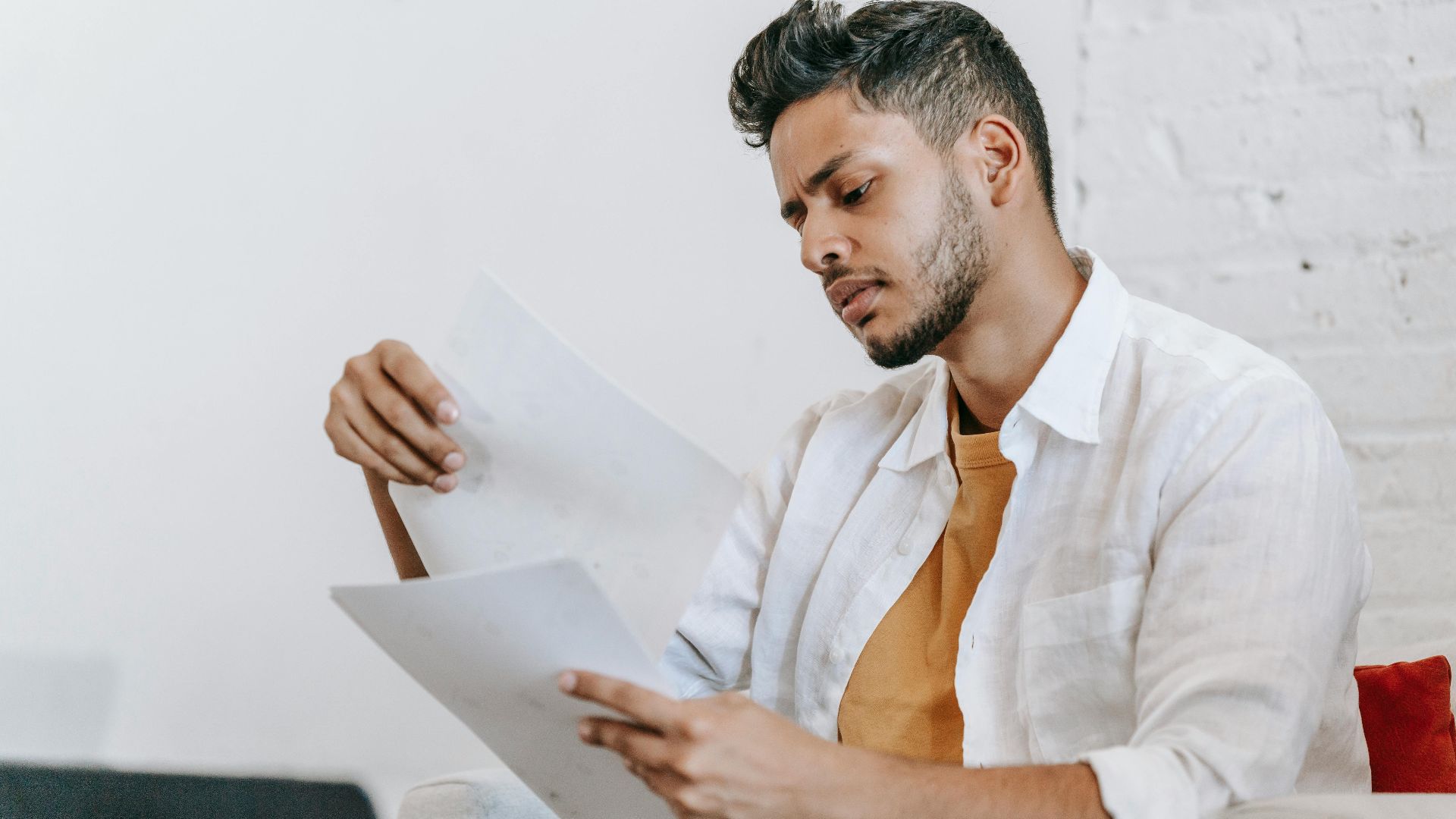 Concentrated ethnic male in casual wear reading documents and sitting on armchair near opened netbook
