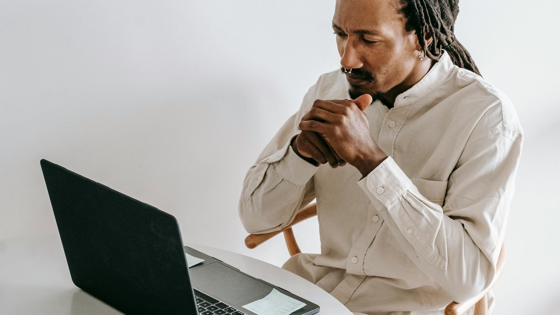 Concentrated adult African American male in formal wear sitting at table with cup of coffee and watching video on netbook in light room