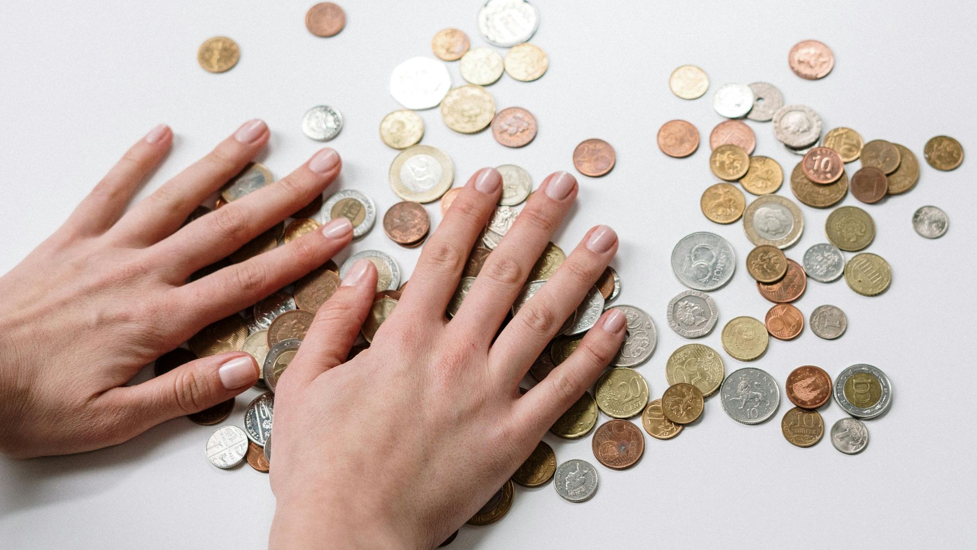 Hands arranging a variety of international coins on a neutral light background.