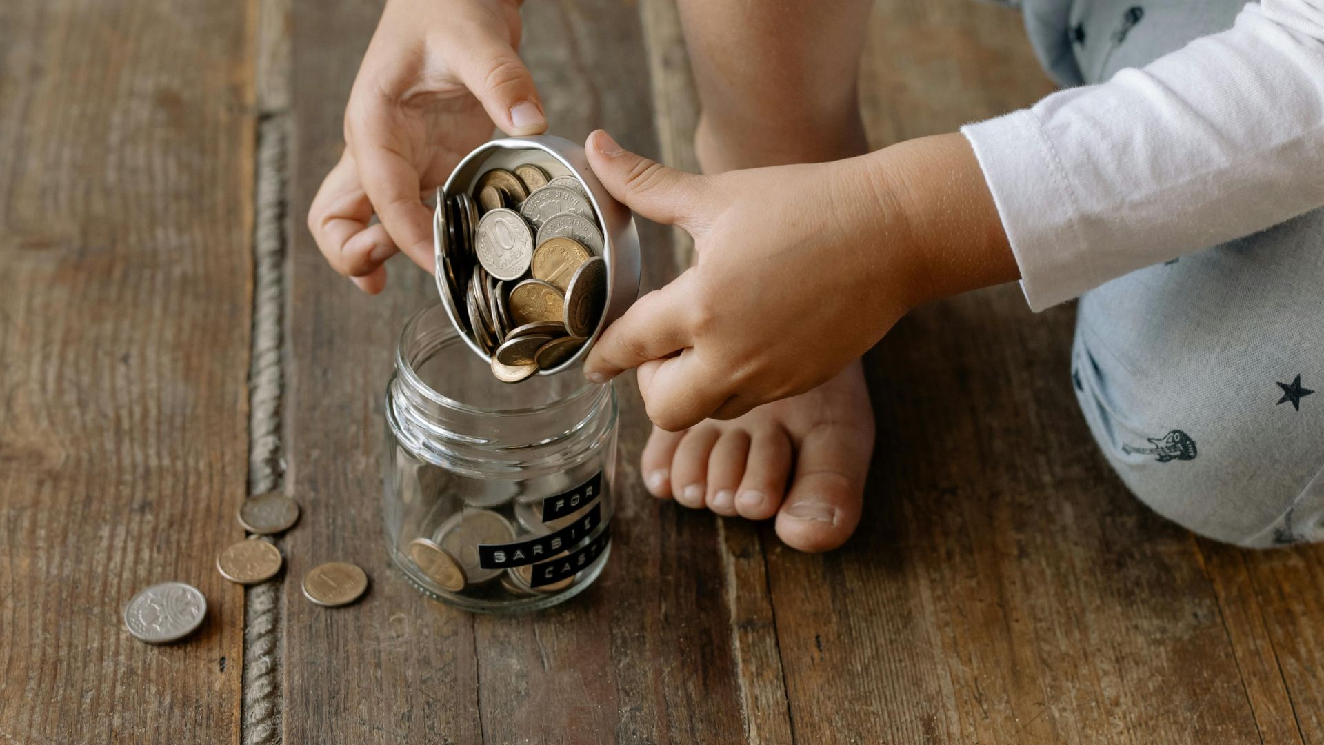 A child adds coins into a glass jar labeled for savings on a wooden floor.