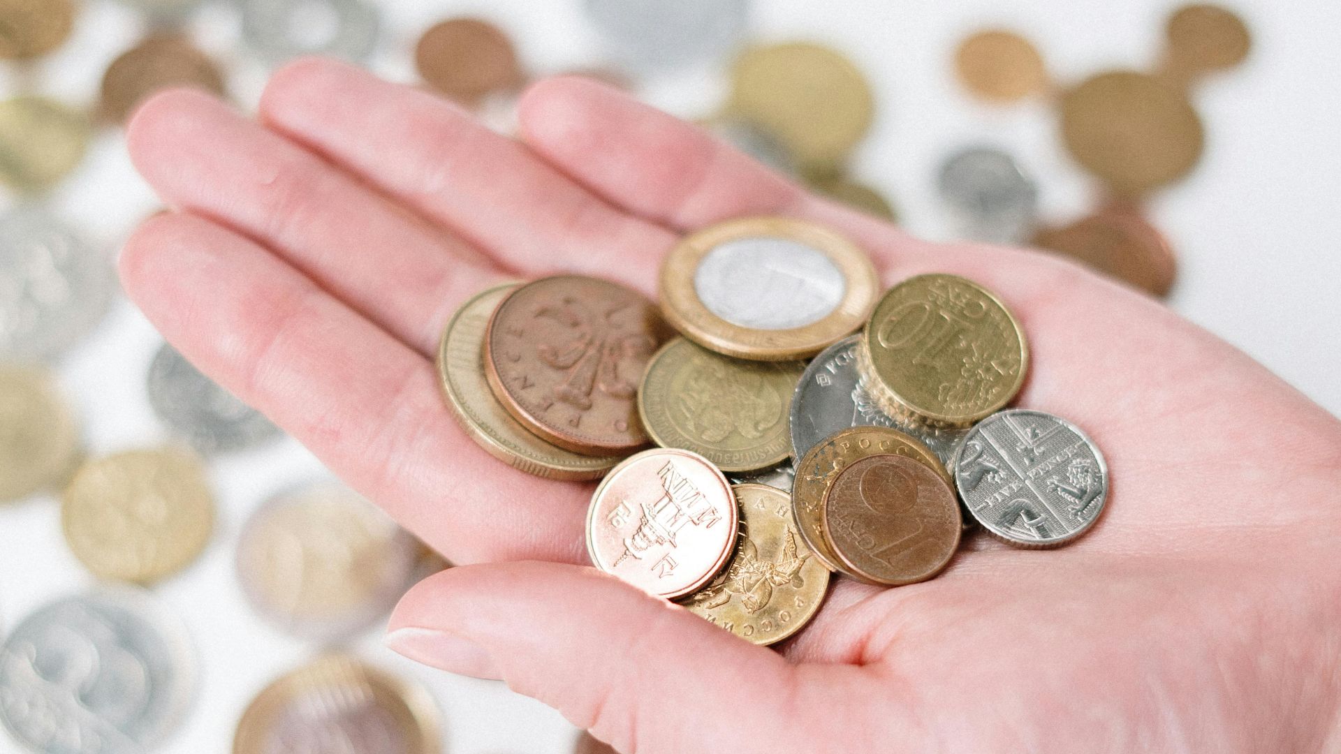A hand holding a variety of coins with more scattered on a white surface, symbolizing currency and finance.