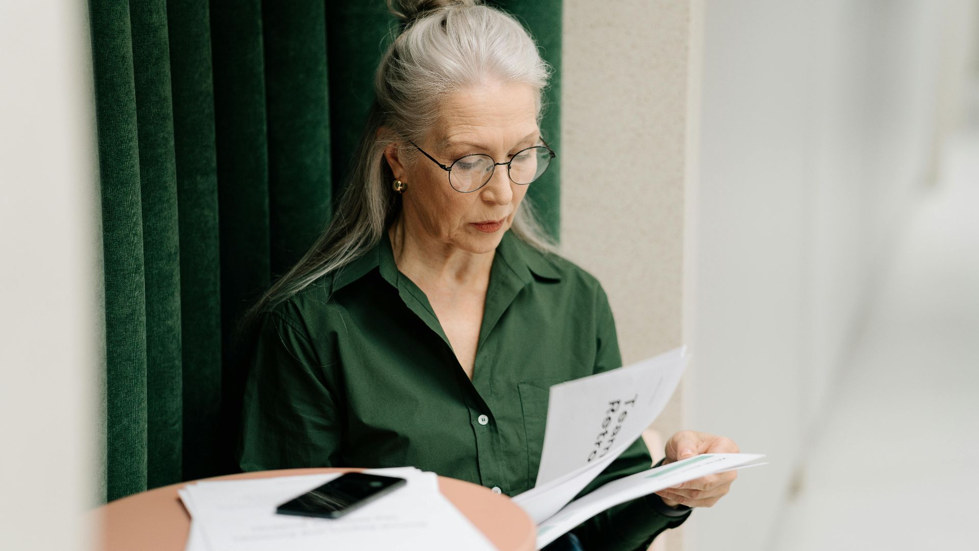 Elderly woman with gray hair reading papers at a small table indoors, focused and thoughtful.