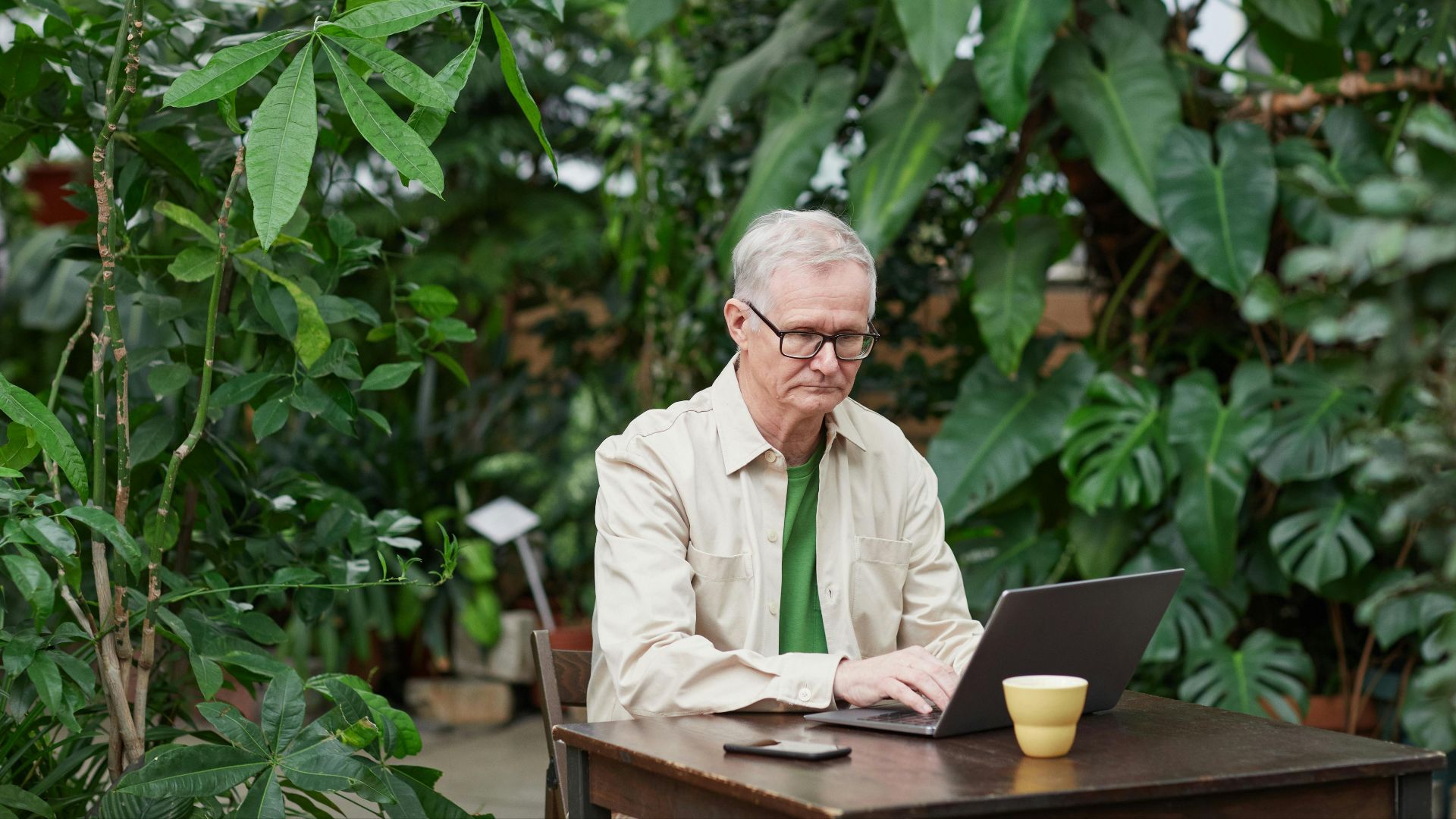 Elder man using laptop in a lush outdoor setting, showcasing freelance remote work lifestyle.