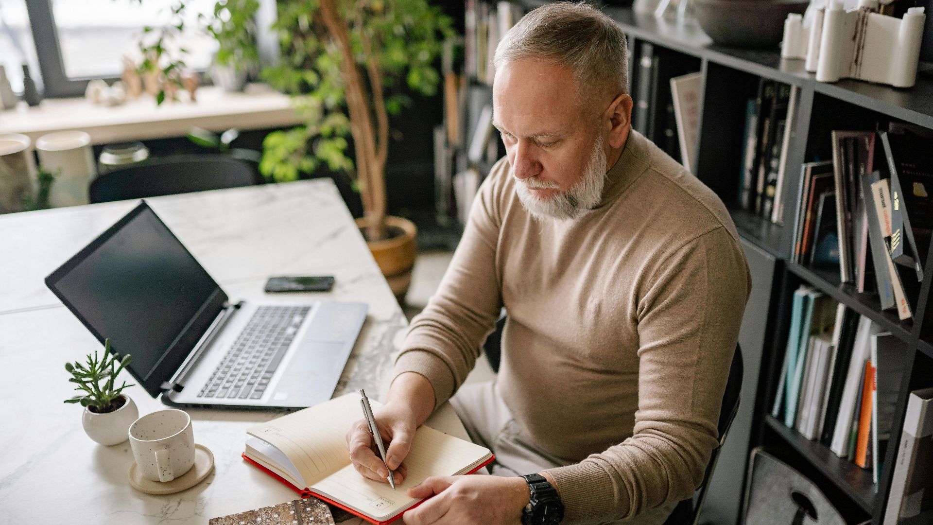 A mature man with a beard writes in a diary at his home office desk next to a laptop.