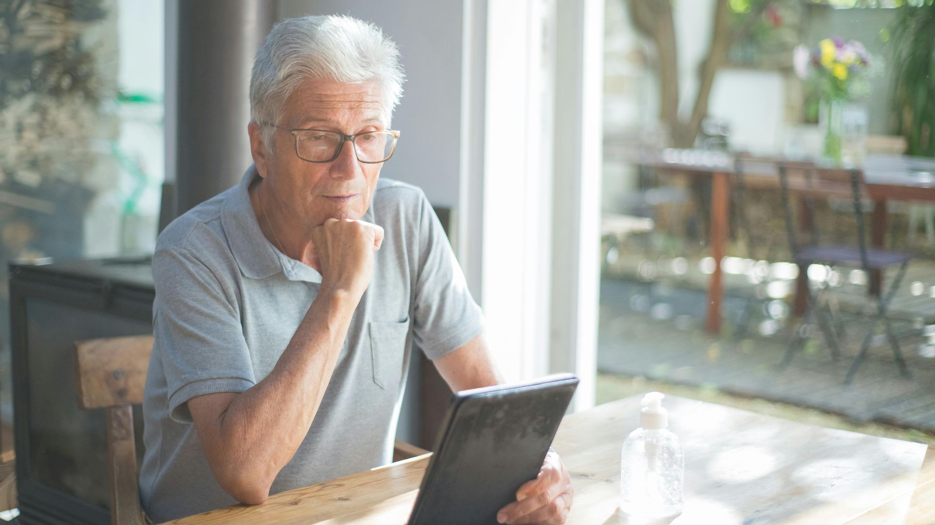 Senior man with eyeglasses reading a tablet at home in a sunlit room.