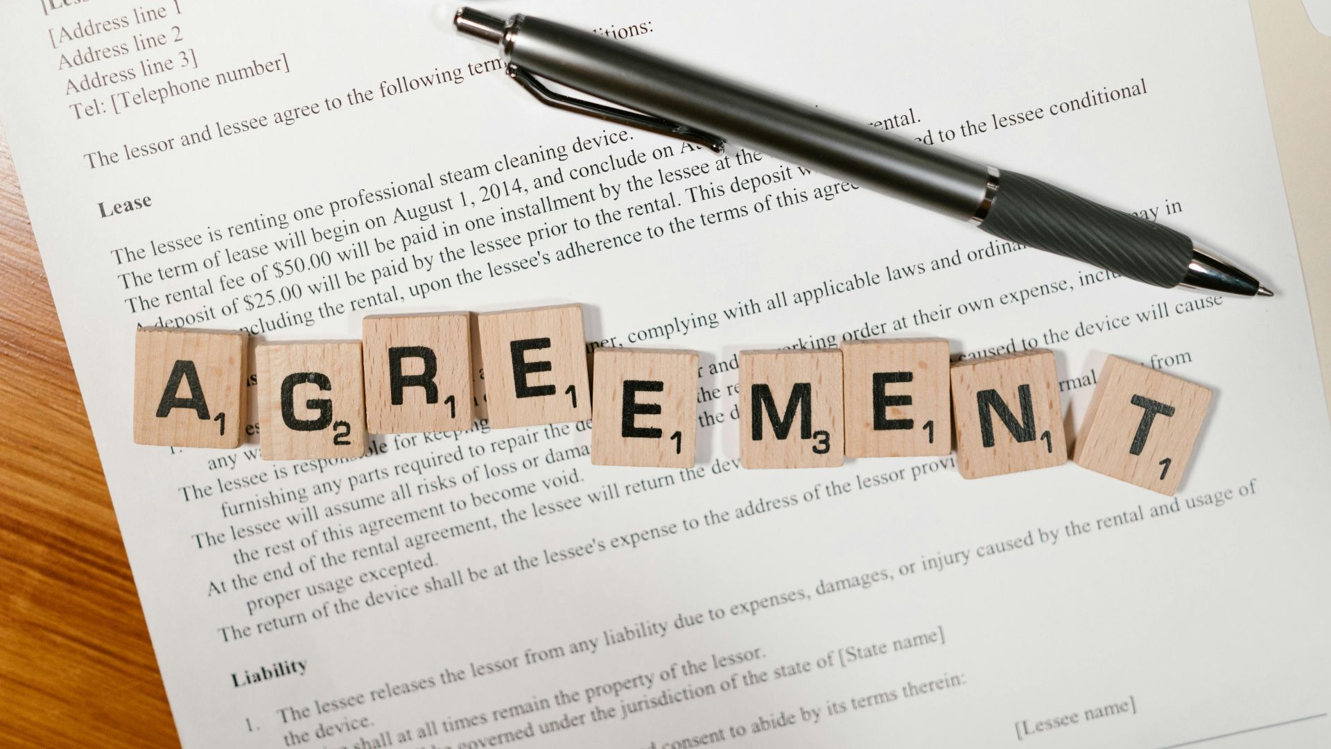 A document with Scrabble tiles spelling 'AGREEMENT' and two pens, symbolizing contract signing.