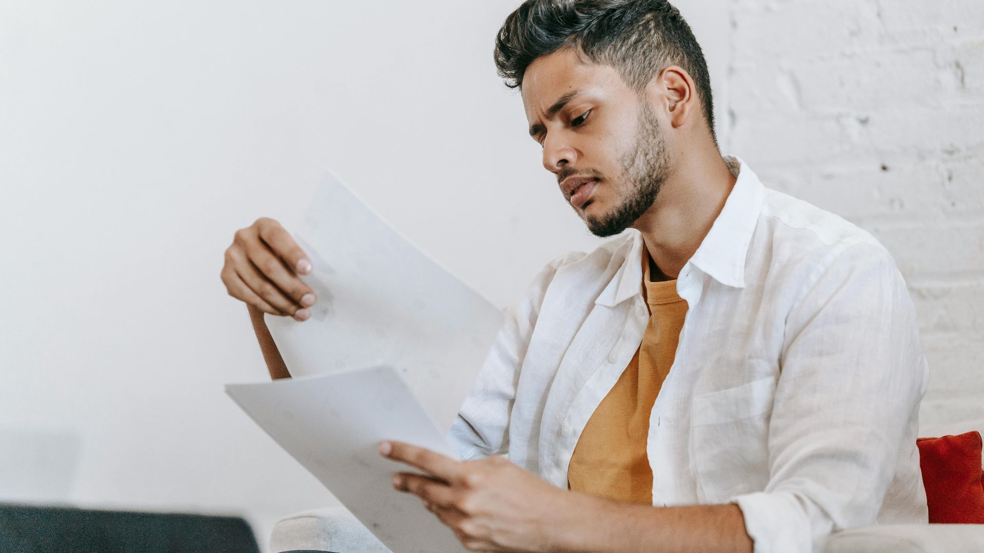 Concentrated ethnic male in casual wear reading documents and sitting on armchair near opened netbook