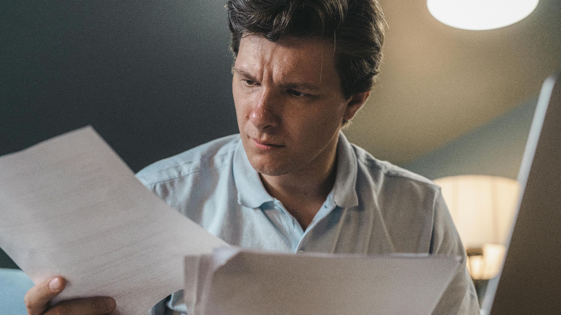 A man intently examines papers, seated indoors under warm lighting, focusing on his work.