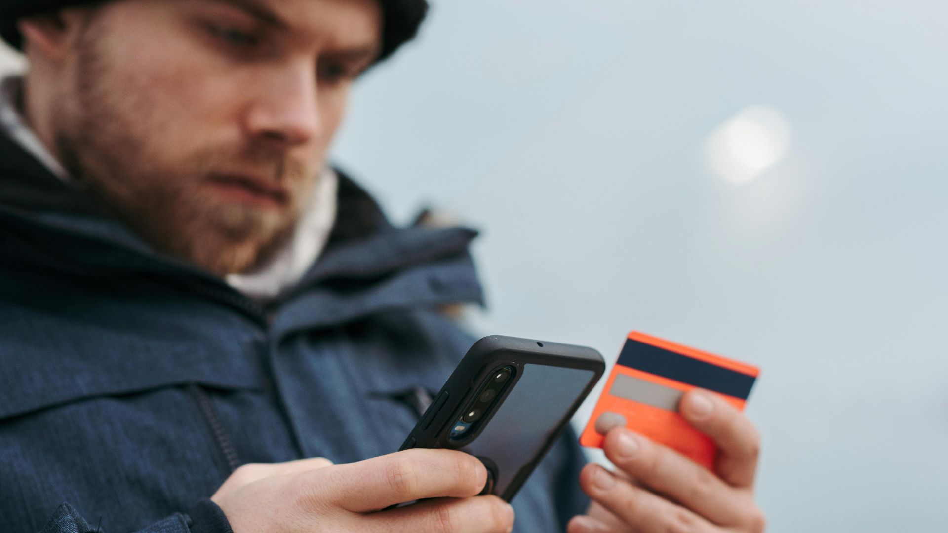 Crop male in outwear entering details of credit card on mobile phone while making online payment for purchase in daytime