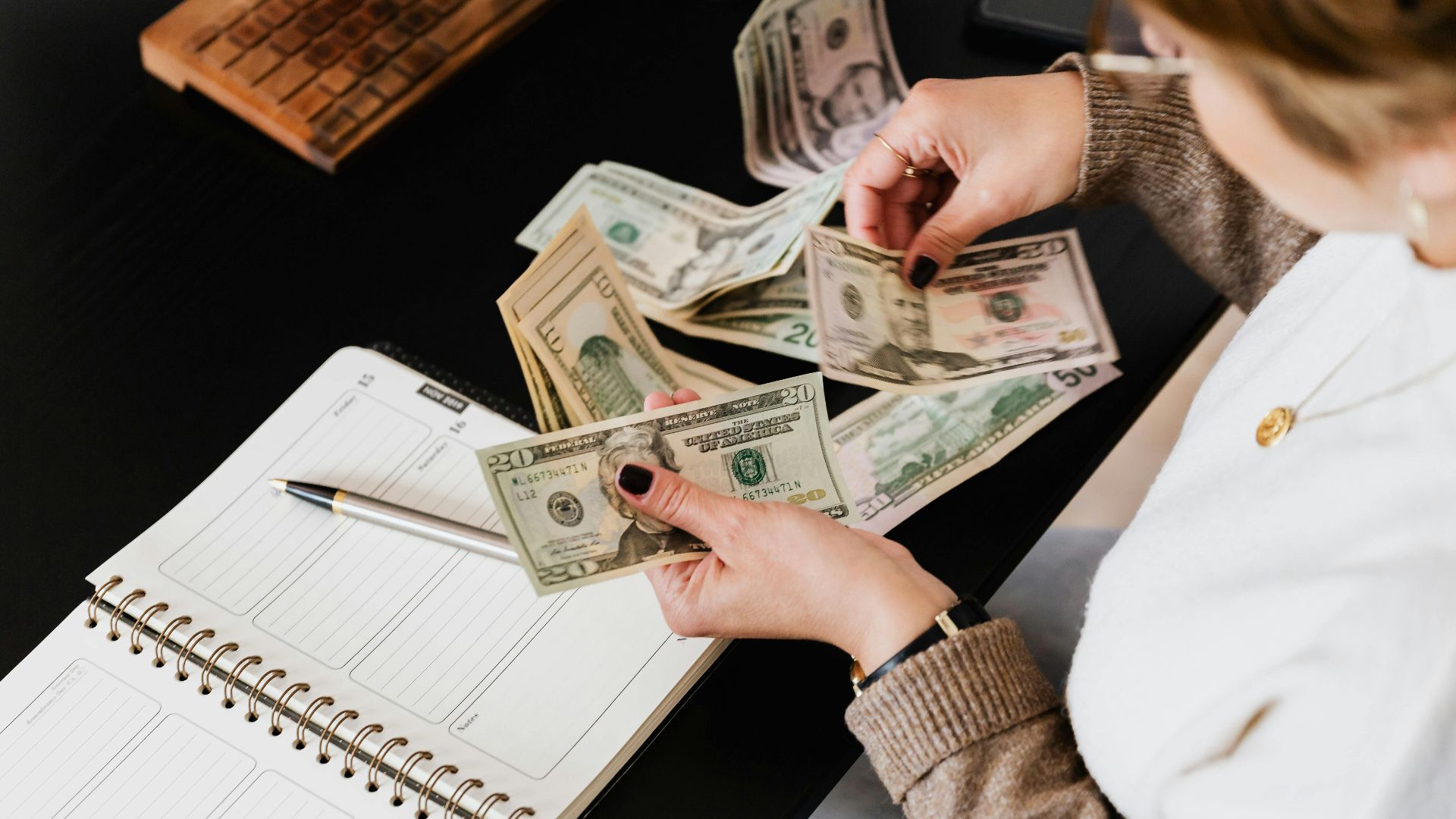 Woman organizing finances, counting dollar bills at desk with open notebook.