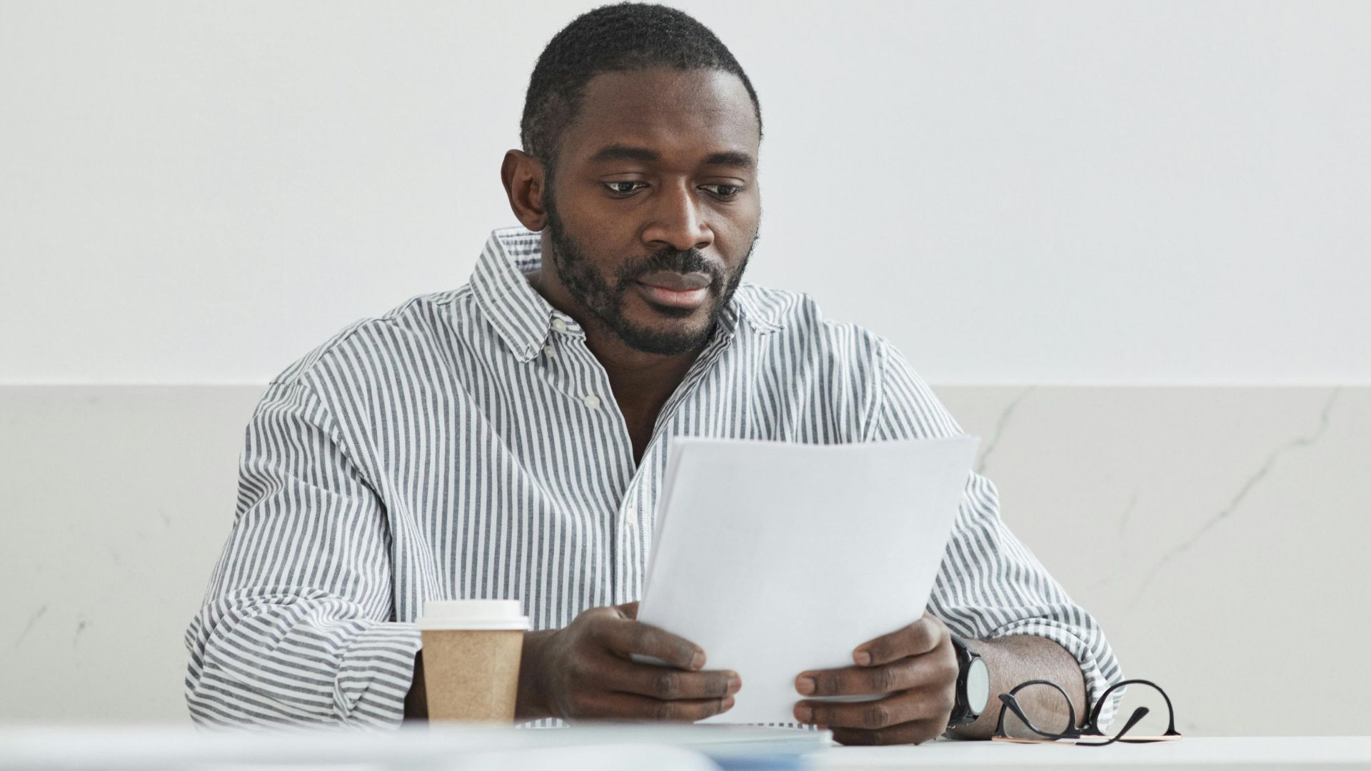African American man sitting indoors, reading papers with a coffee cup nearby.