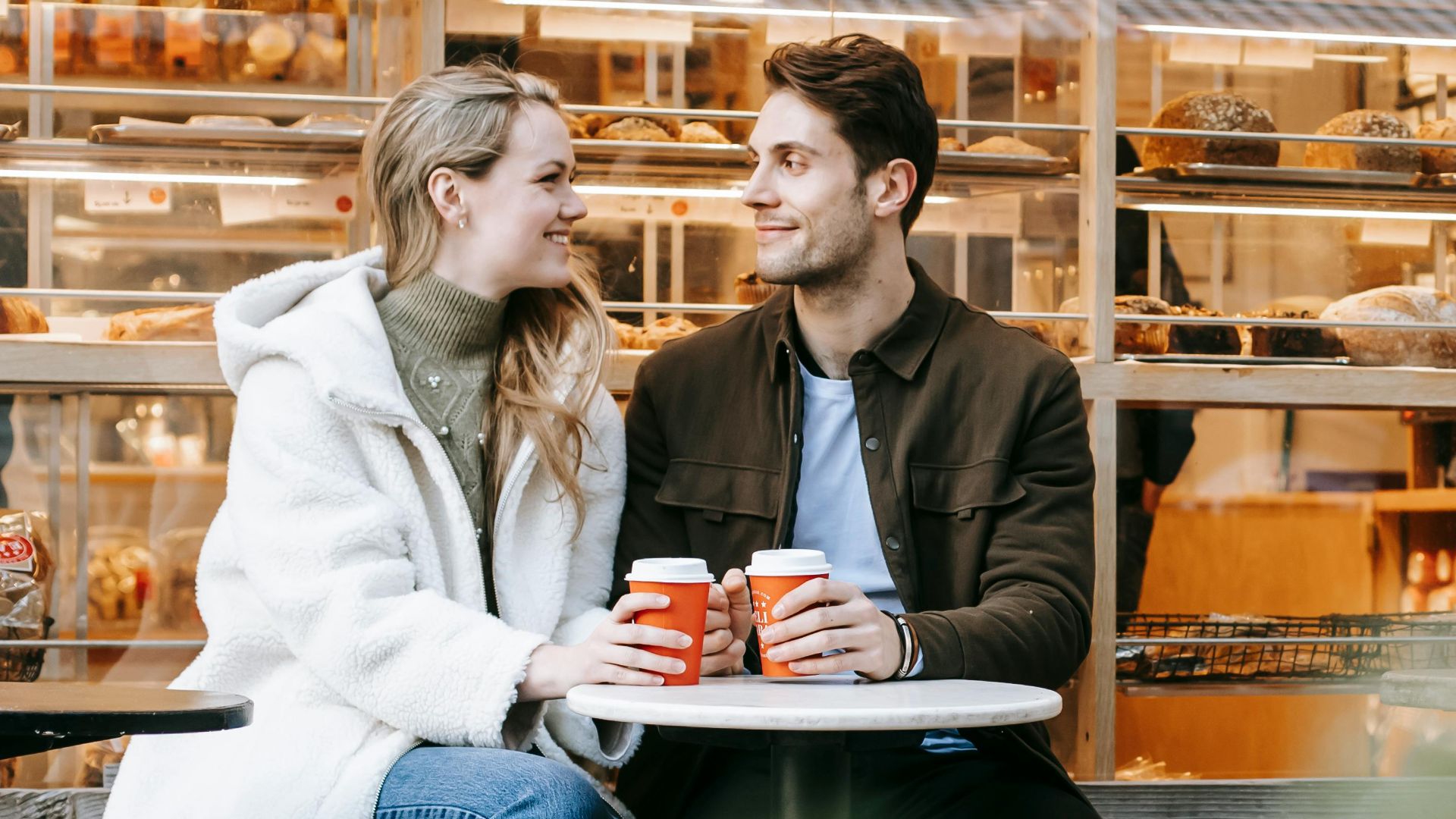 Cheerful young couple enjoying hot drink and looking at each other in small bakery
