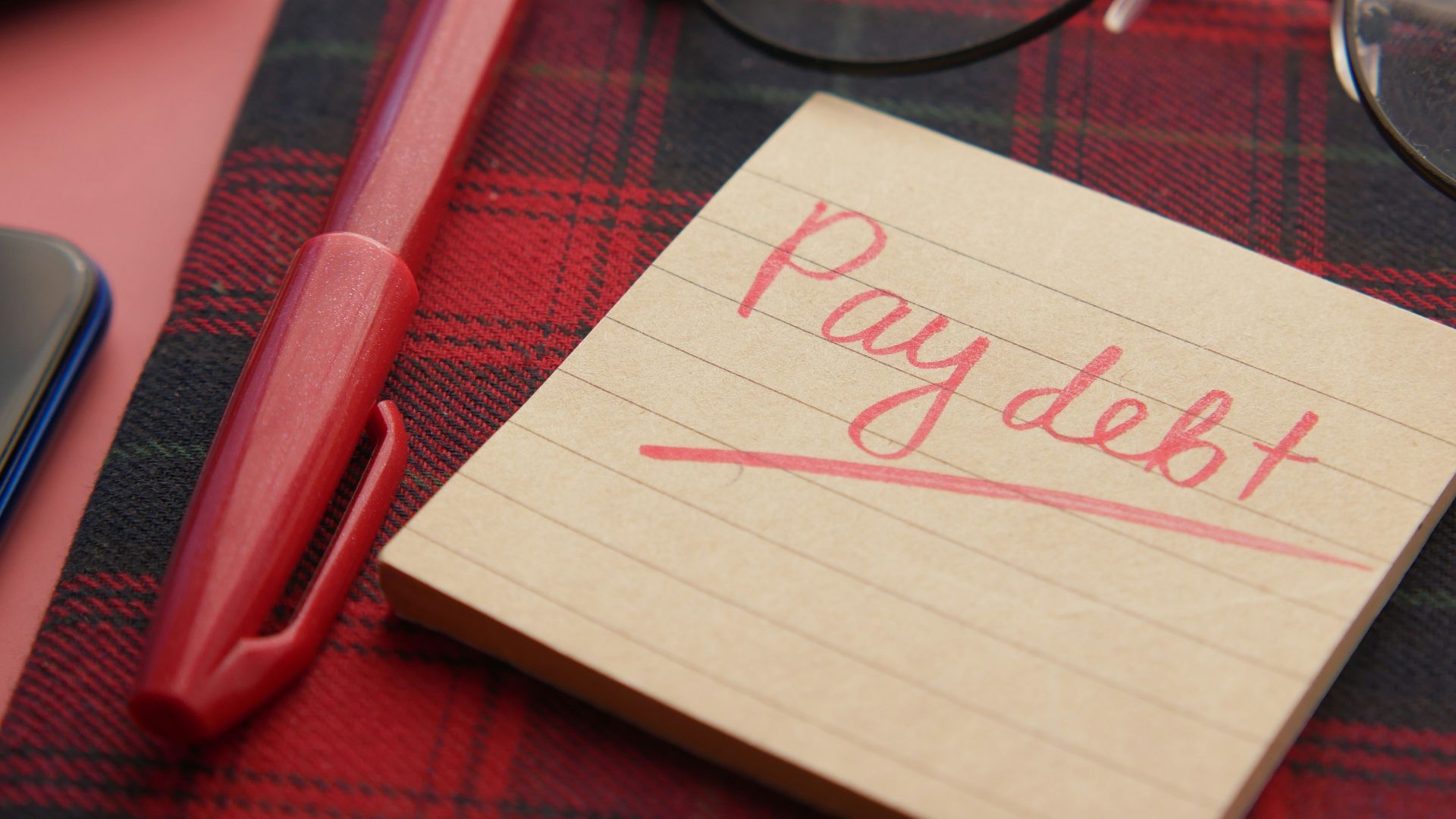 a note that says pay debt next to a pen and glasses