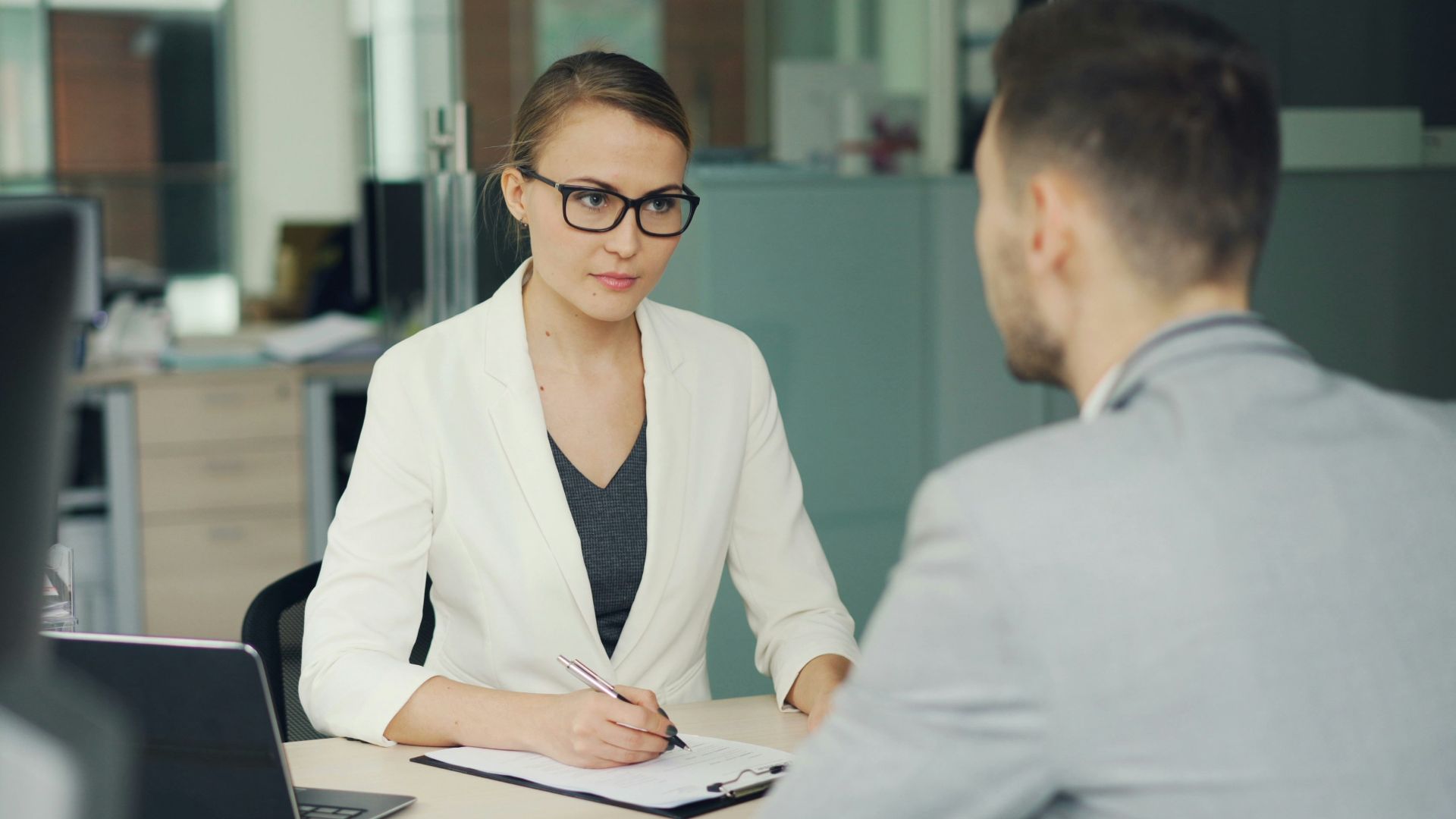 Woman in glasses interviews man at office desk.