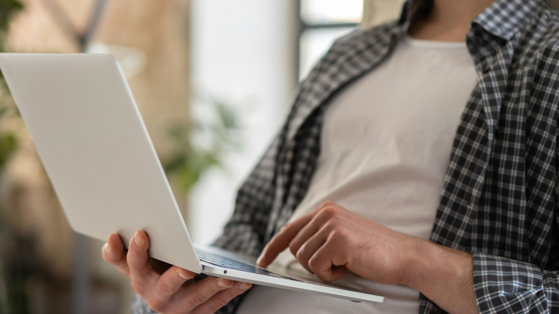 Young adult in casual shirt using a laptop indoors with blurred background and shallow focus.