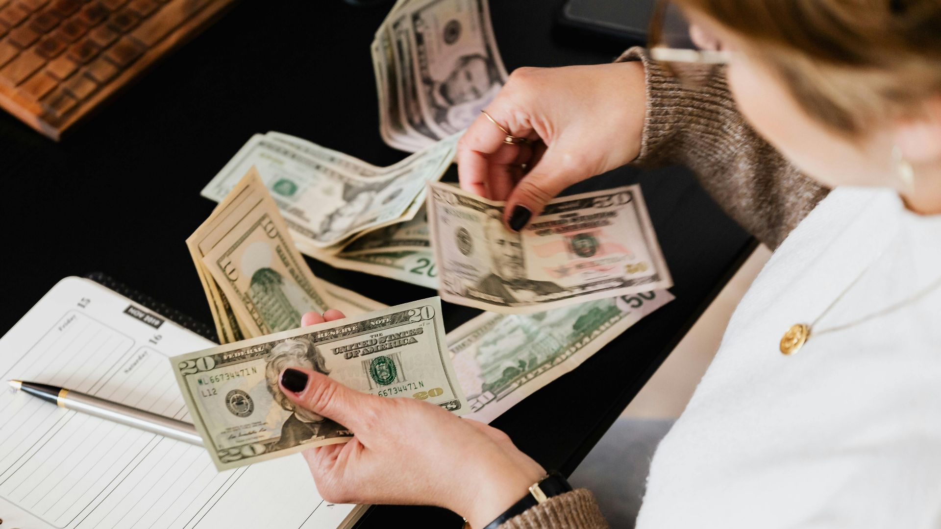 Woman organizing finances, counting dollar bills at desk with open notebook.