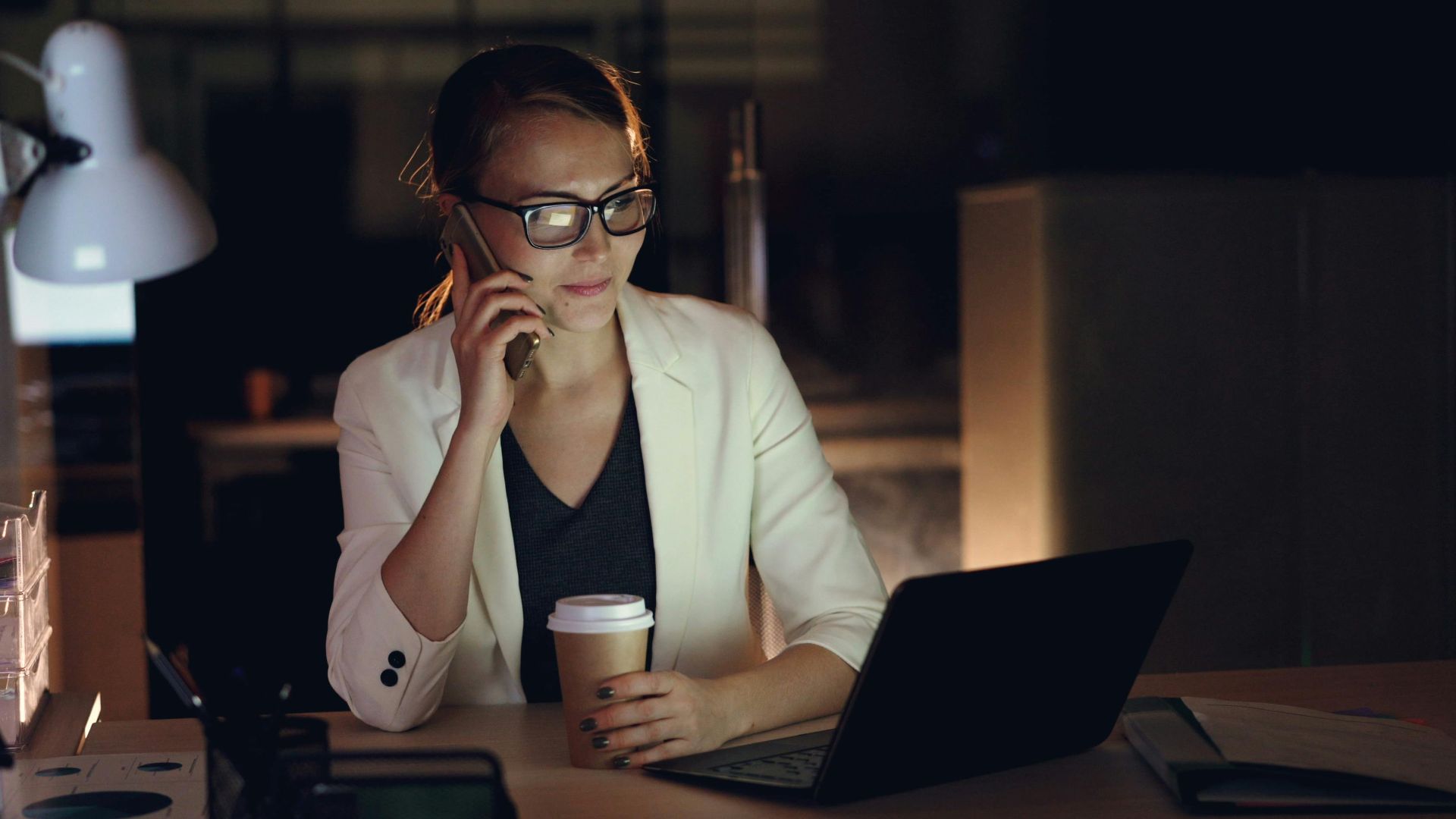 Woman in glasses working late at night on laptop, holding coffee, making phone call.