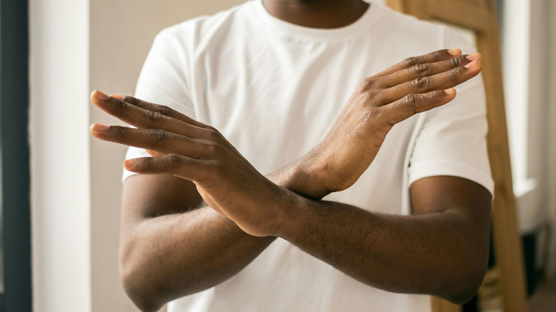 Crop unrecognizable bearded African American male in white t shirt demonstrating no gesture with hands while standing in light room