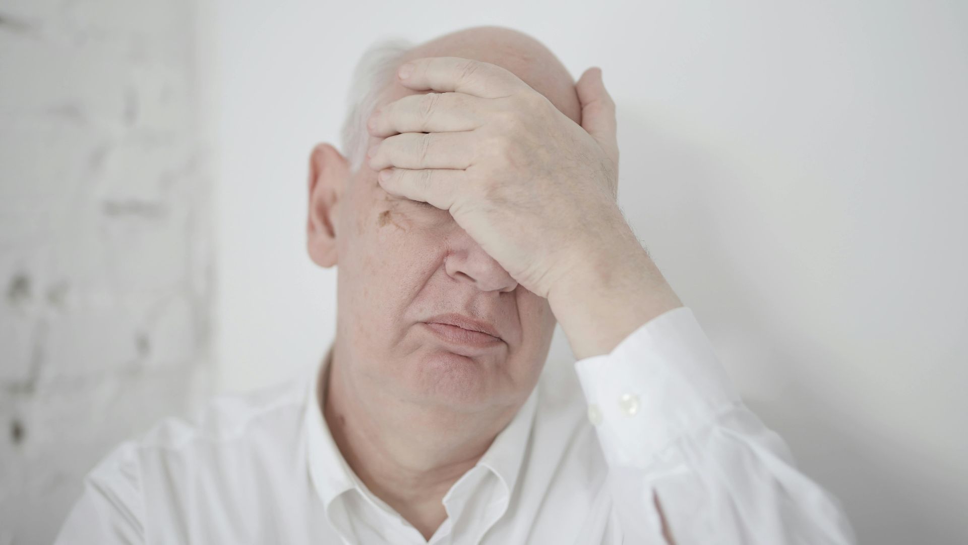 Disappointed senior bald man in white shirt covering face with hand while sitting near wall in light room