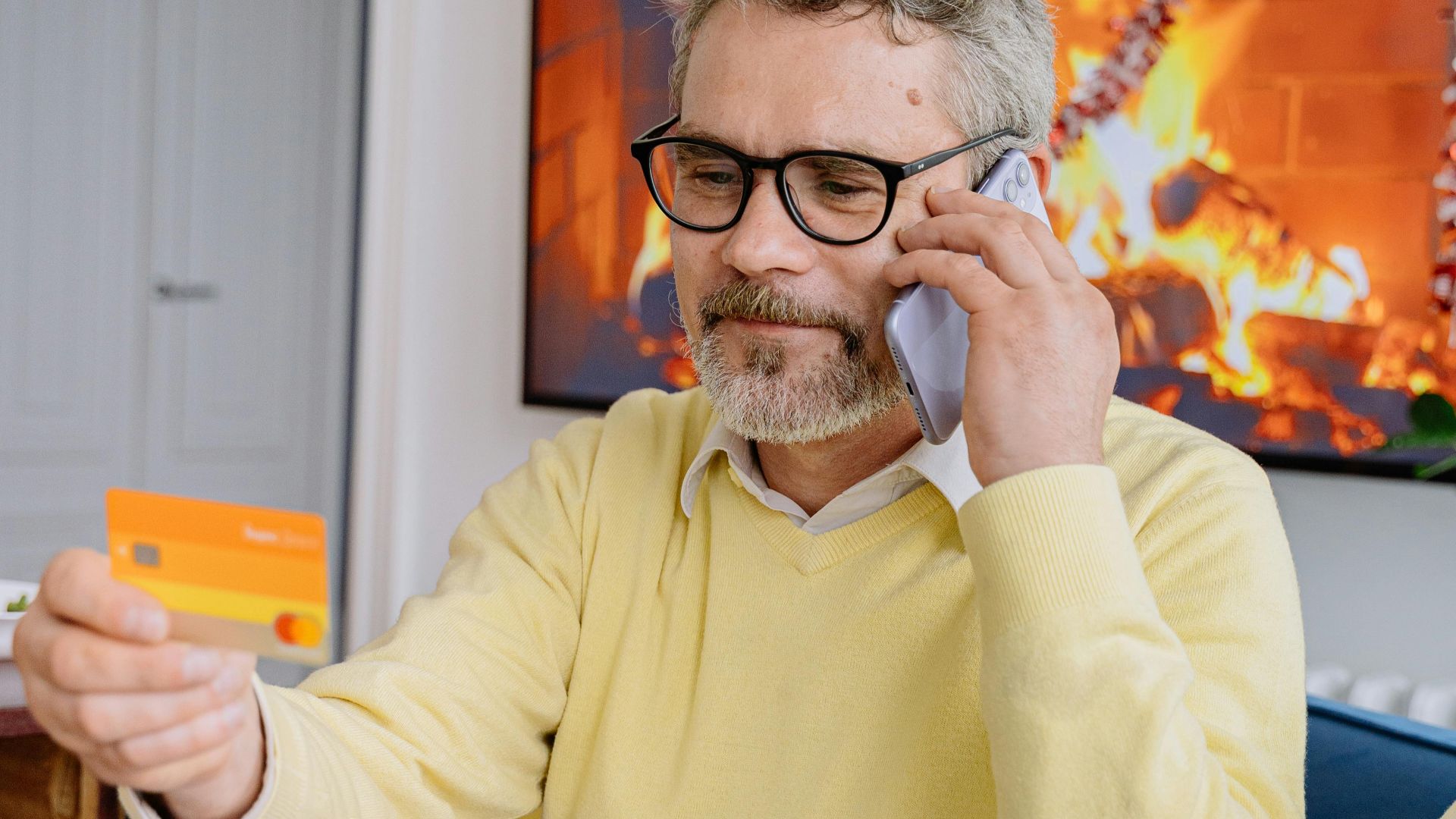 Elderly man in yellow sweater using phone and credit card while sitting indoors by a fireplace.
