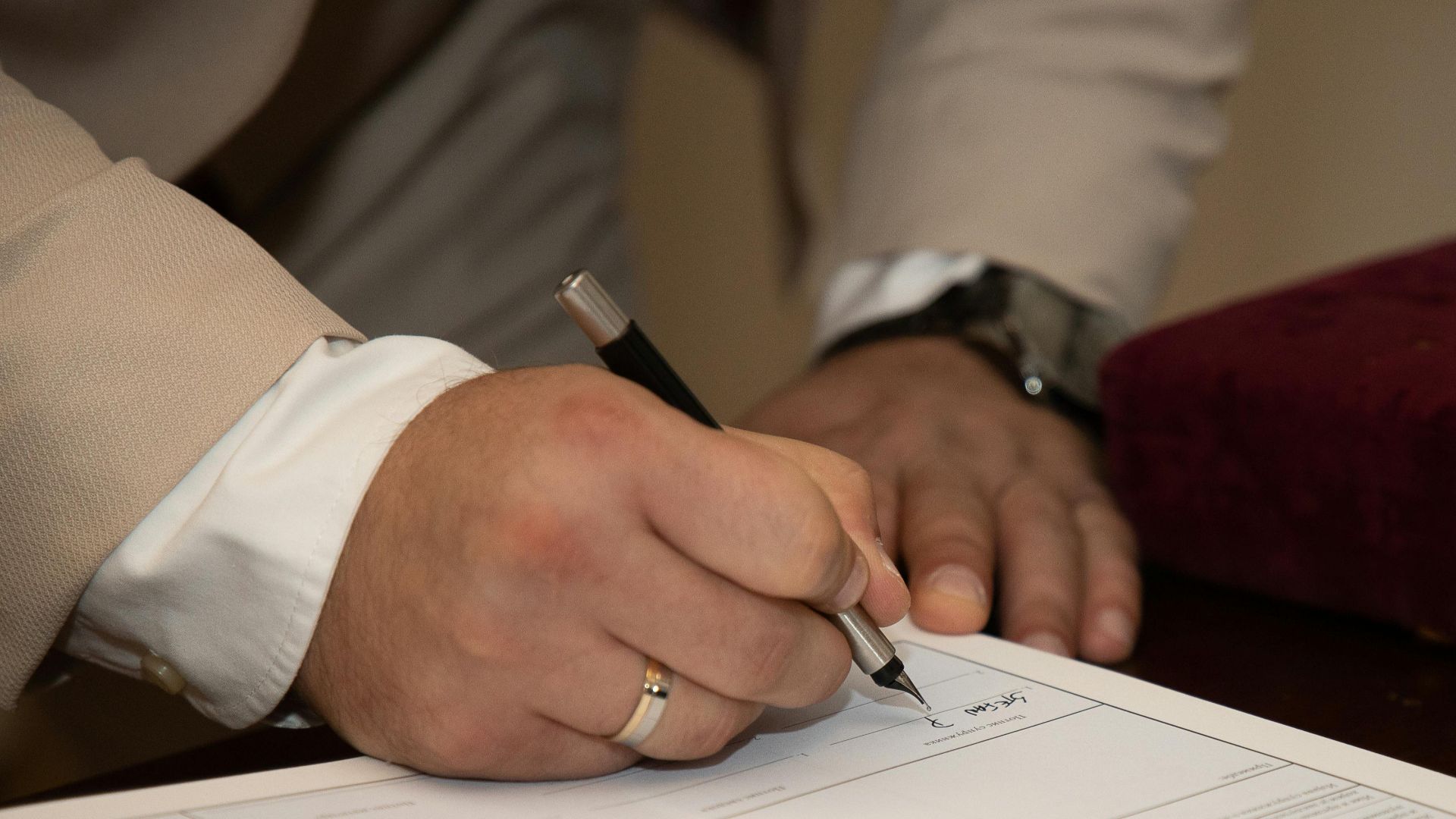 A close-up of a man signing a document, showcasing a wedding ring and pen.