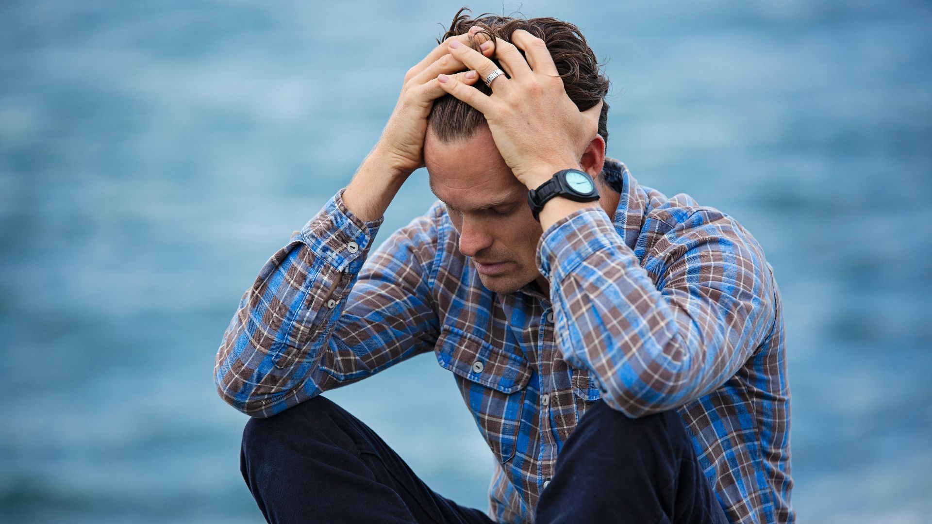 A man in a plaid shirt sits by the water looking distressed, symbolizing stress.