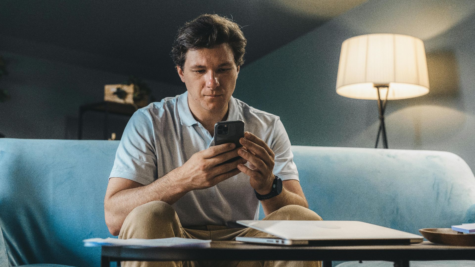 A man sitting on a sofa indoors using a smartphone under warm lighting.