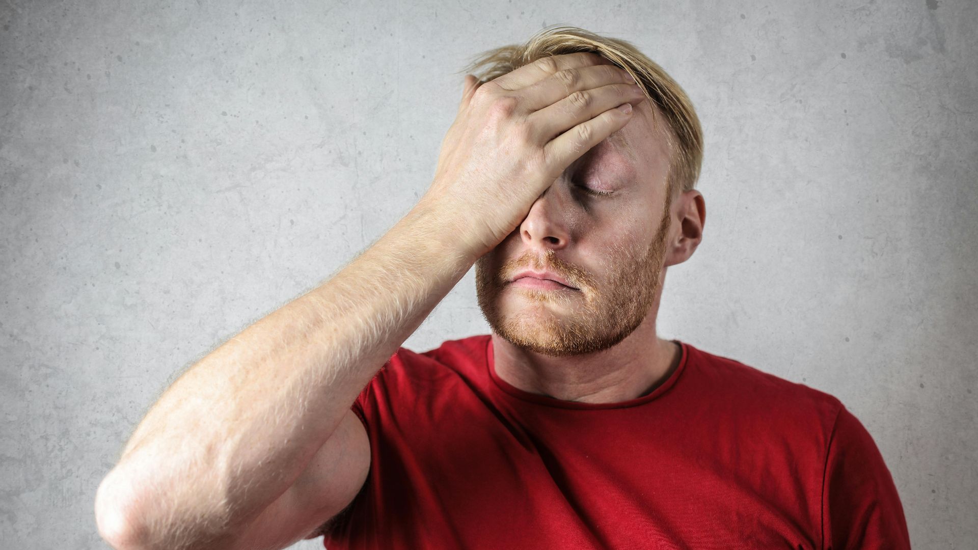 A frustrated man in a red shirt holds his head in stress against a neutral background.
