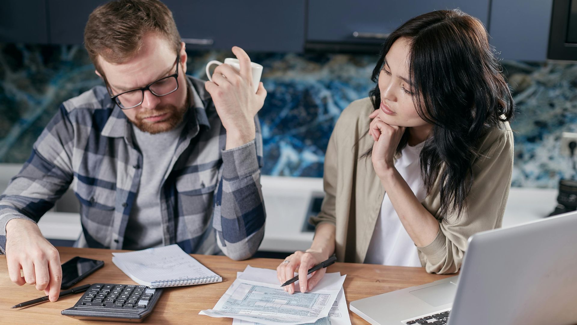 A couple reviews bills and documents on a laptop while discussing household finances.