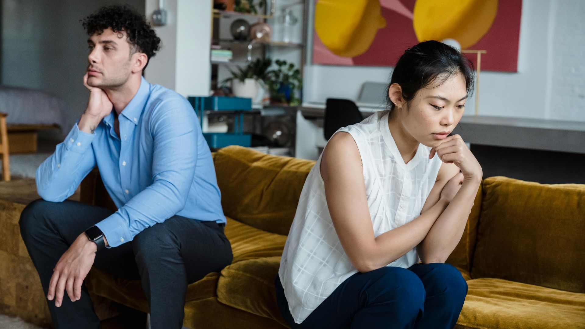 A couple sits indoors on a sofa, both appearing thoughtful and contemplative.