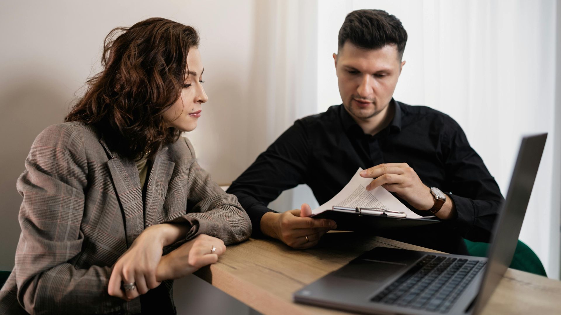 Two professionals discussing documents at a desk with a laptop, focused on collaboration.