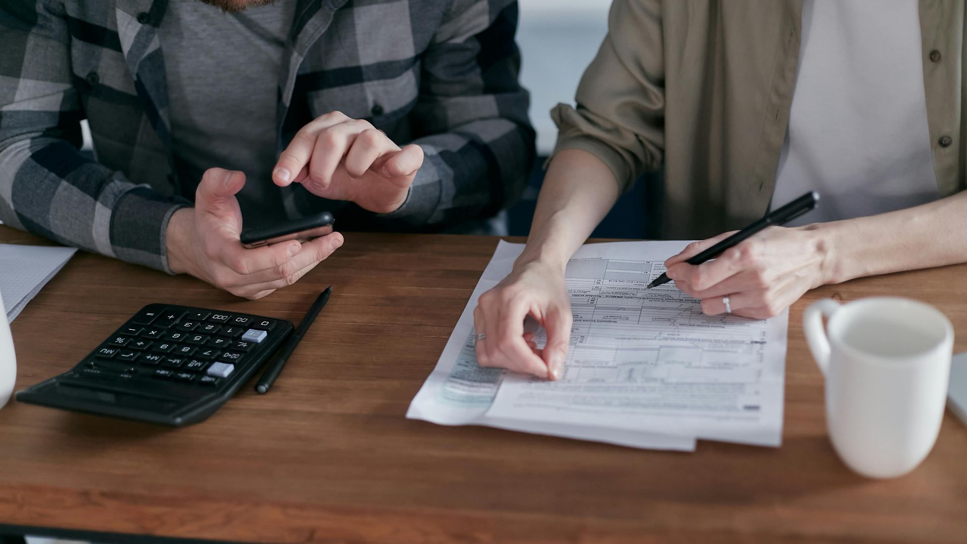 Two people working together on tax forms using a calculator at a wooden desk.