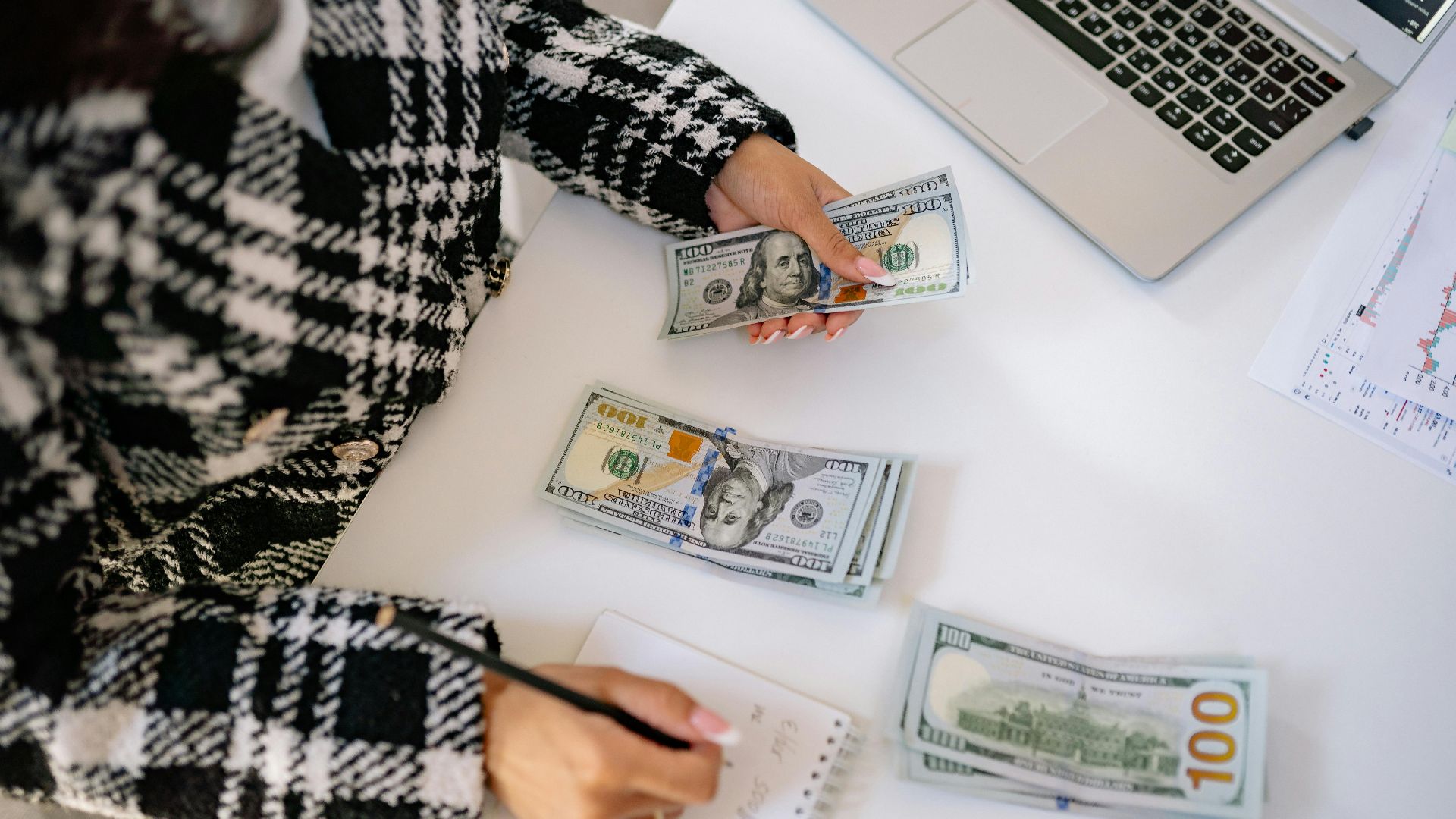 A woman in a plaid coat counts American dollars at a desk with a notebook and laptop.