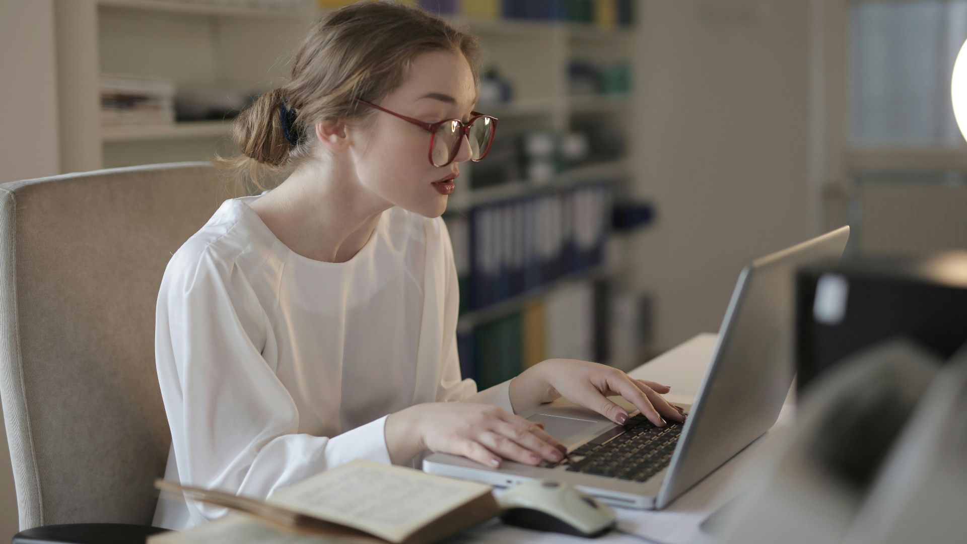 Professional woman in glasses working at a desk with a laptop in an organized modern office.