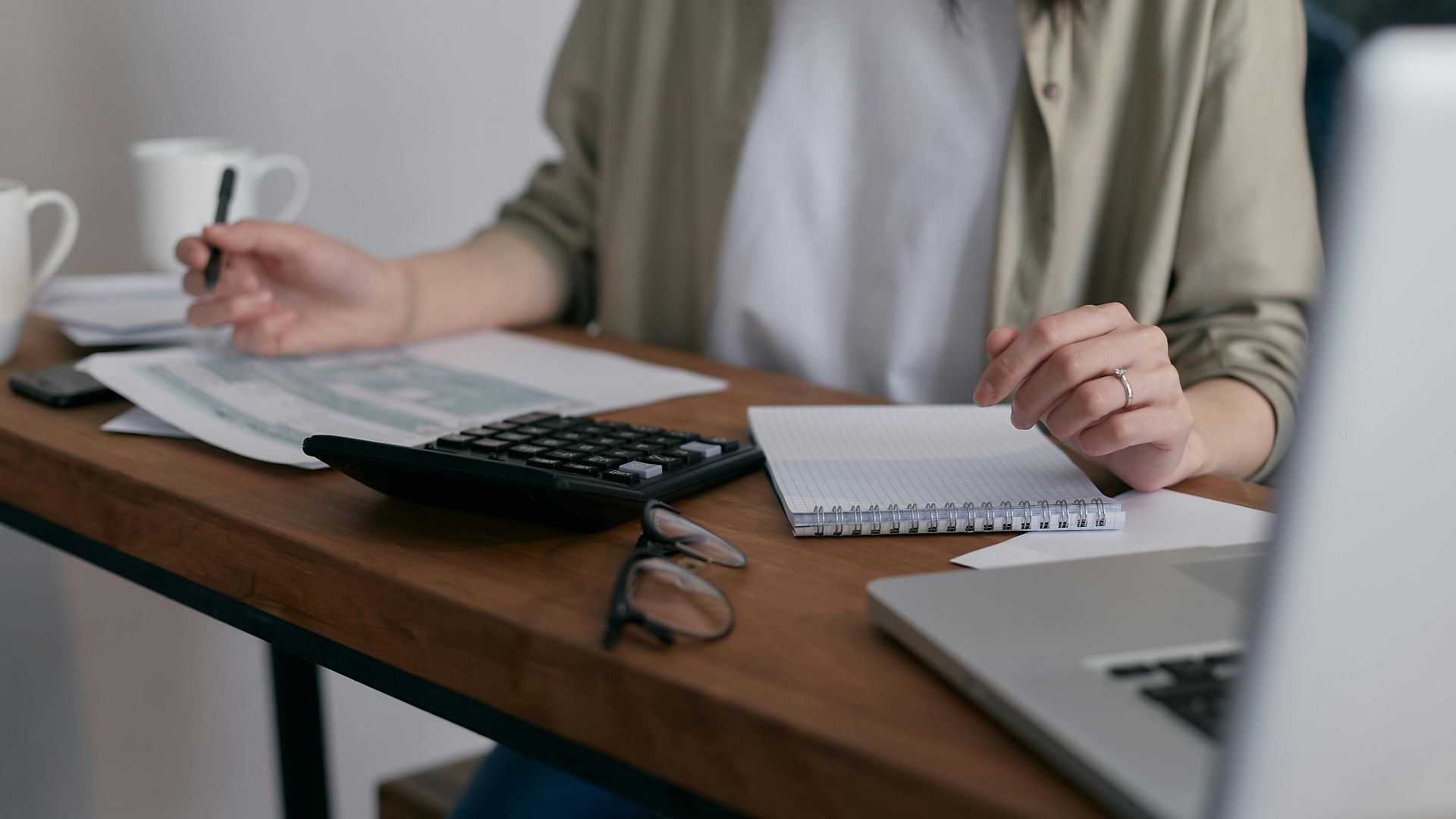 A woman manages finances at home, using a laptop and calculator on a wooden desk.