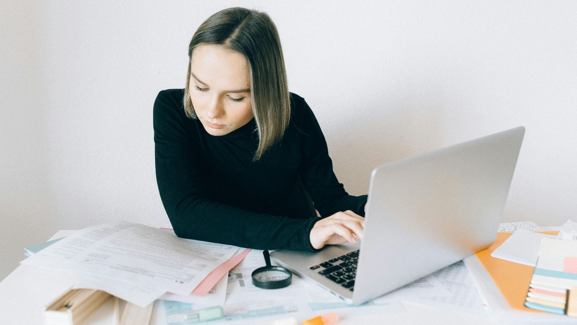 Focused woman researching on a laptop, surrounded by paperwork in a modern office.