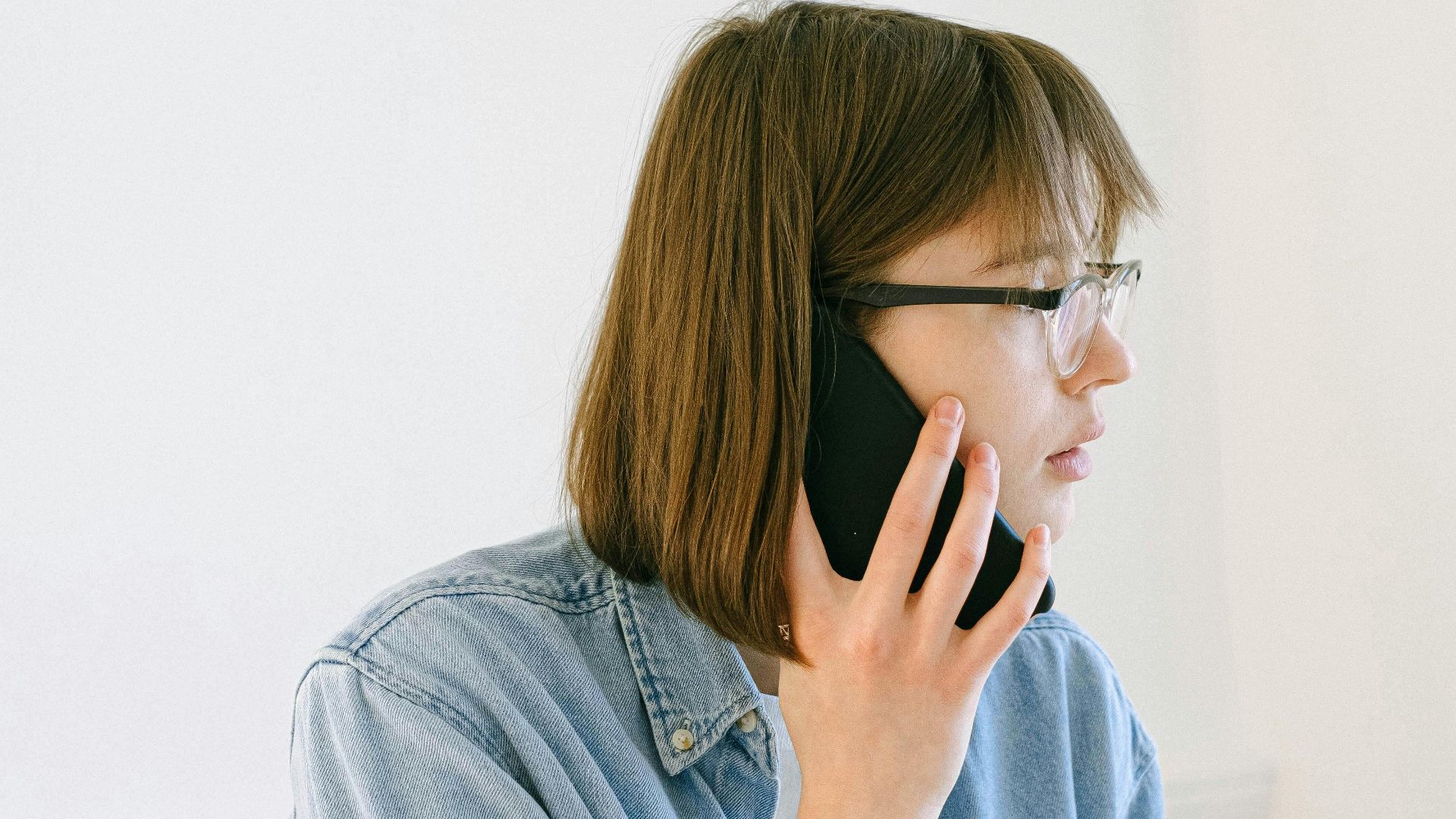A young woman in a denim shirt having a phone conversation indoors during the day.